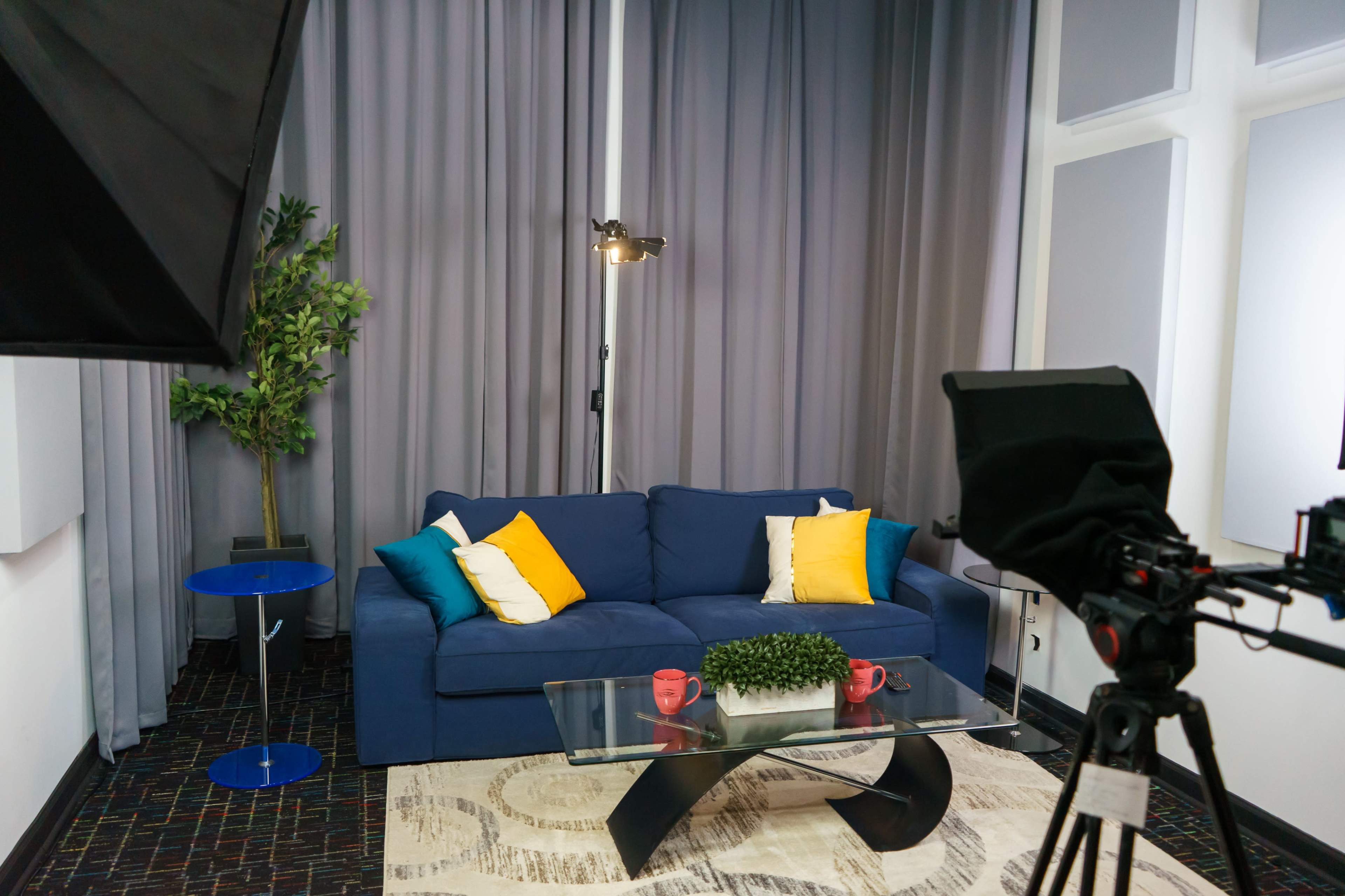 A blue couch with yellow and turquoise pillows is positioned in a room with gray curtains, a glass coffee table, and a camera setup nearby.