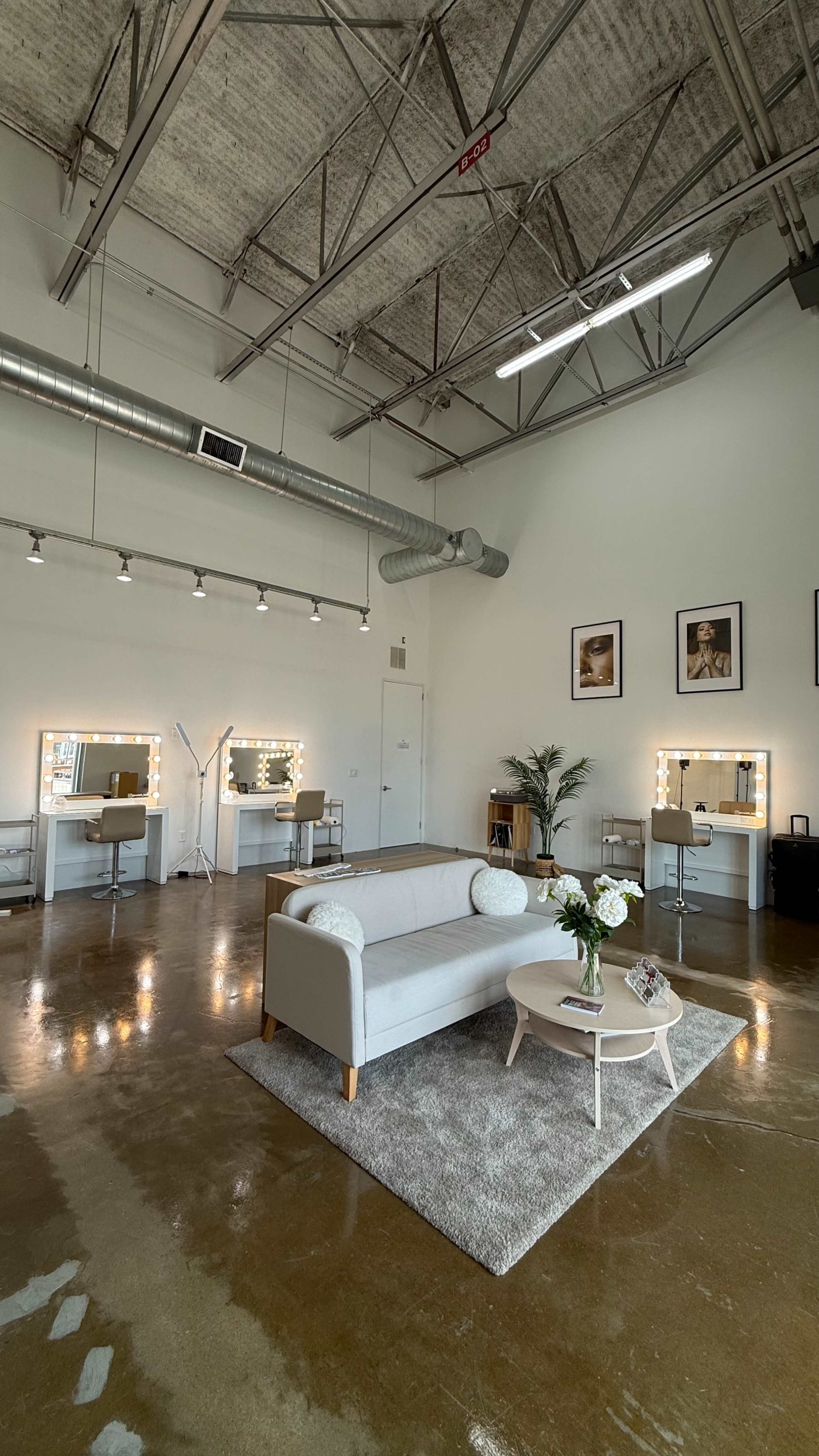 The image shows a spacious, well-lit room with modern furnishings, including a sofa and a coffee table, along with illuminated makeup stations against the walls.