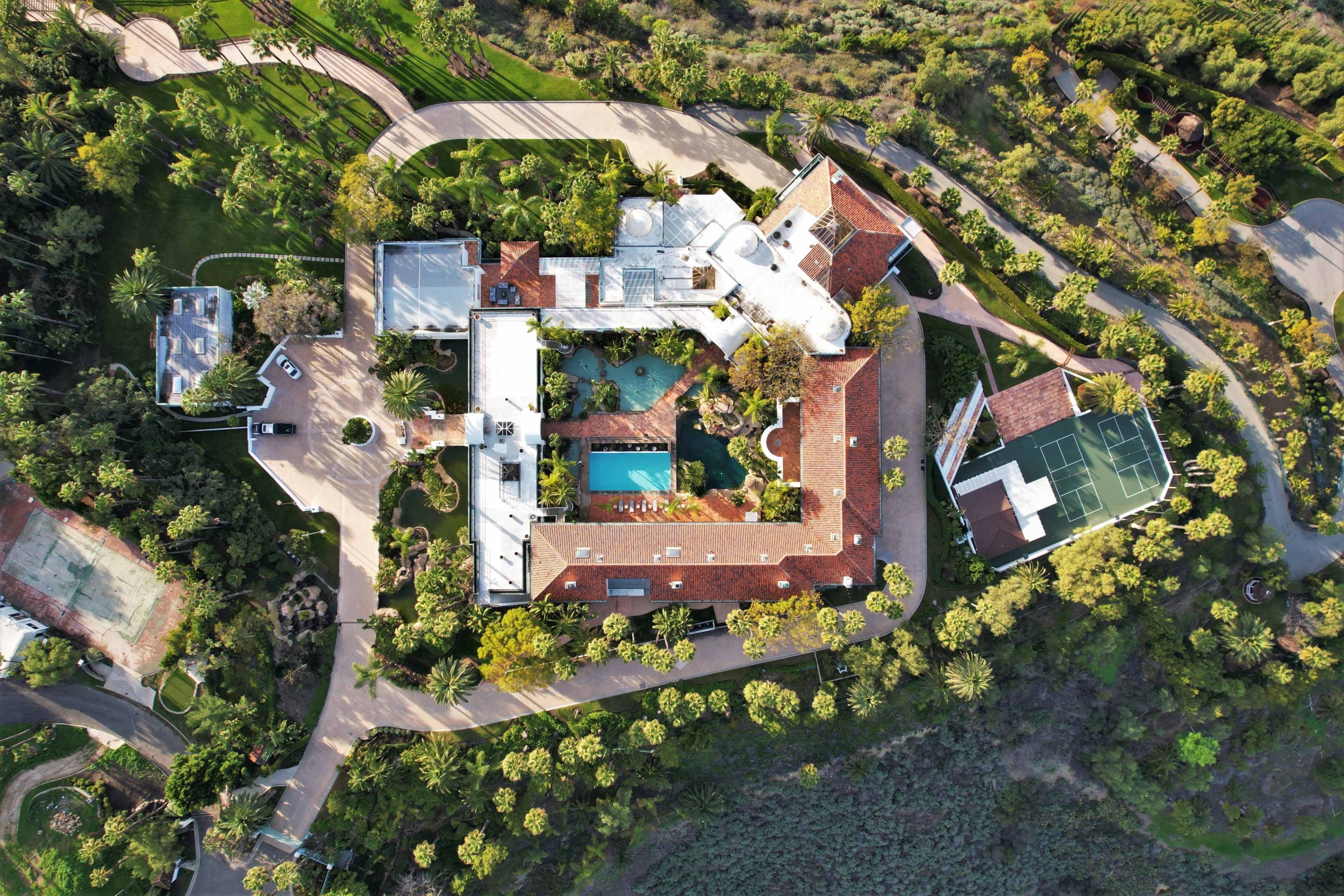 An aerial view of a sprawling estate features a main house with a pool, tennis court, and landscaped gardens surrounded by trees.
