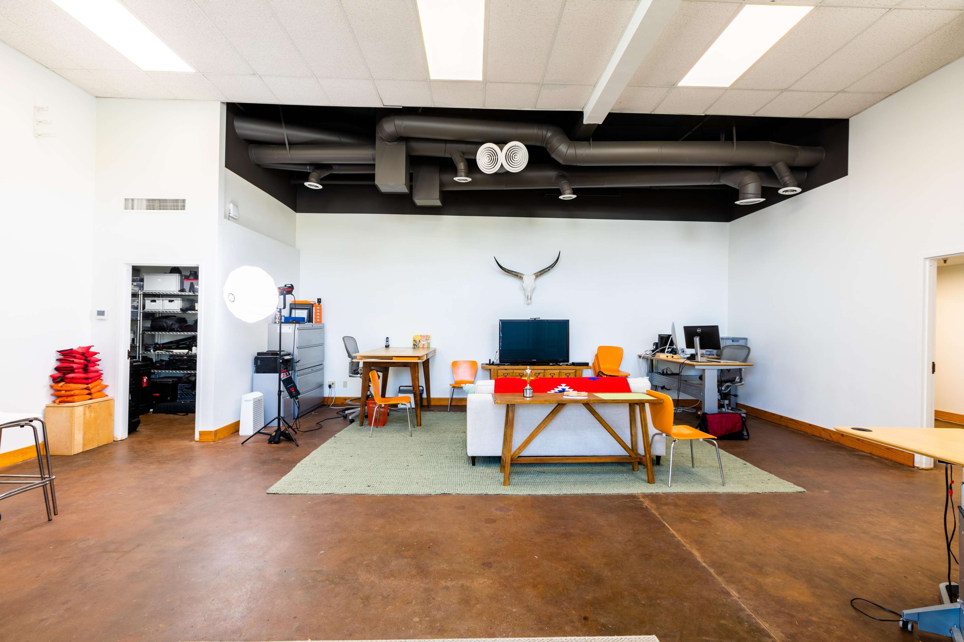 An open office space featuring two wooden tables, orange chairs, and a wall-mounted animal skull, with a large window and exposed ductwork on the ceiling.