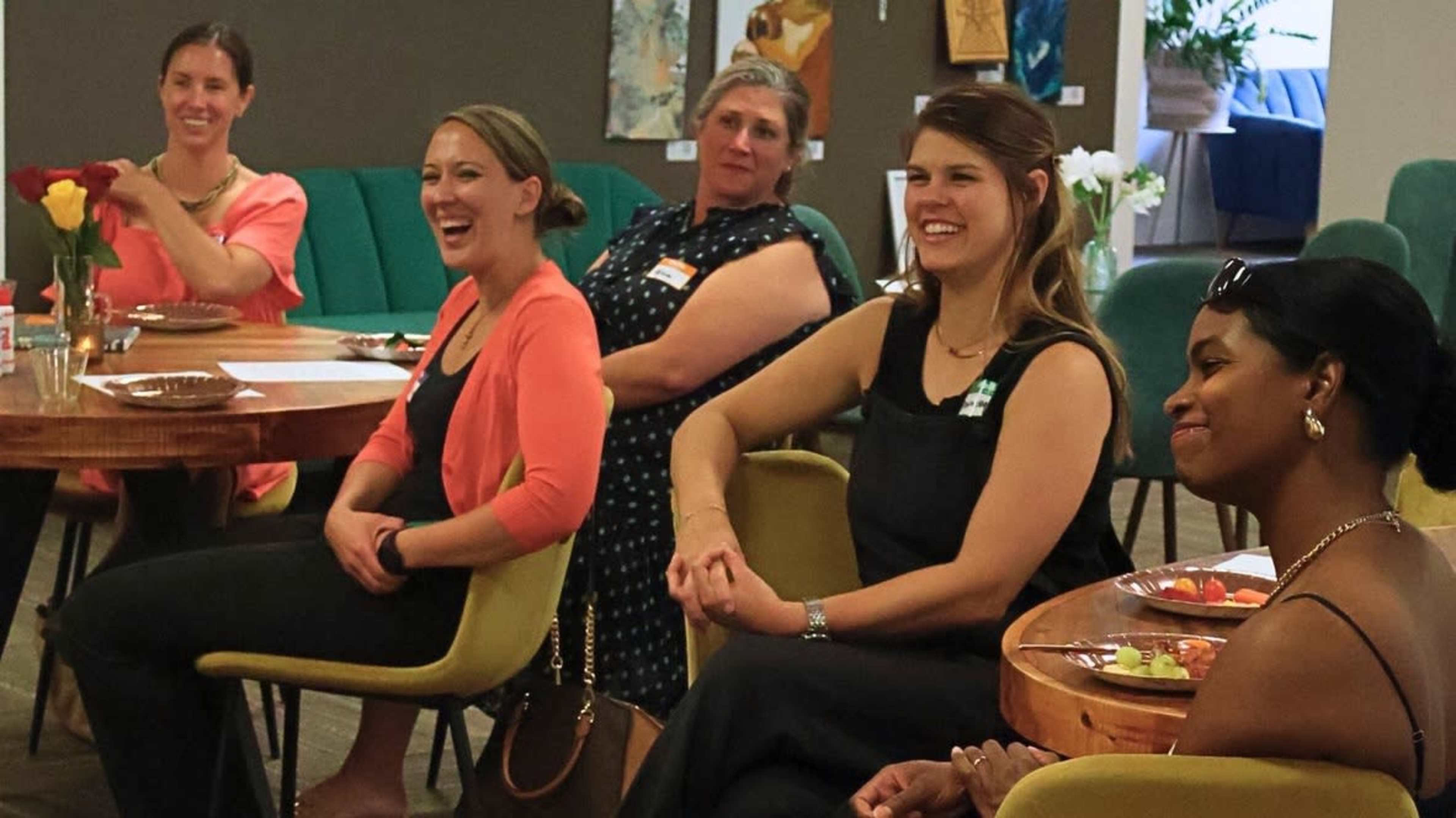 A group of five women sits in a semi-circle, smiling and engaged in conversation, while seated around a wooden table.