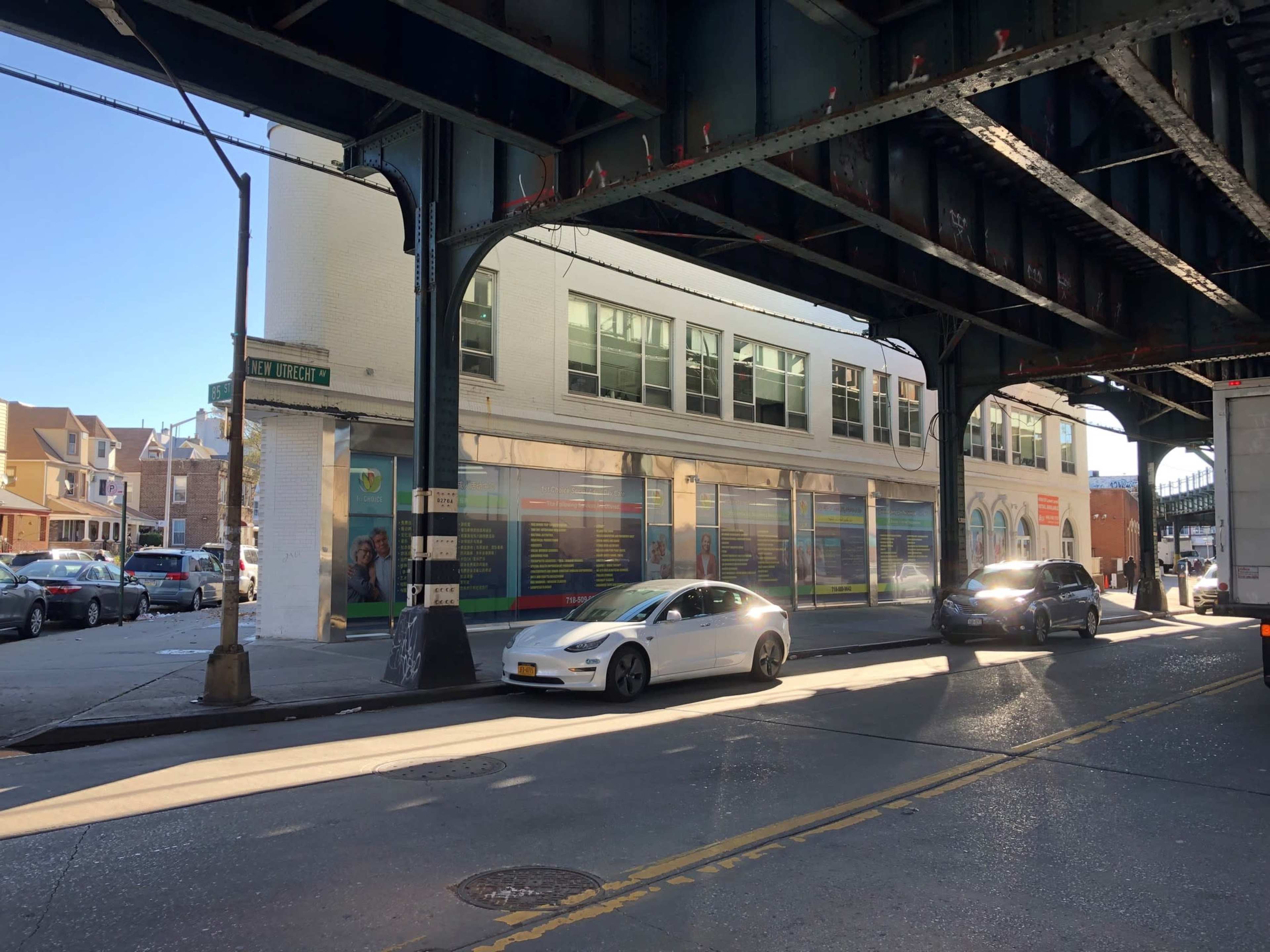 The image shows a street scene with two cars parked on the road under an elevated train track, next to a building displaying colorful advertisements.