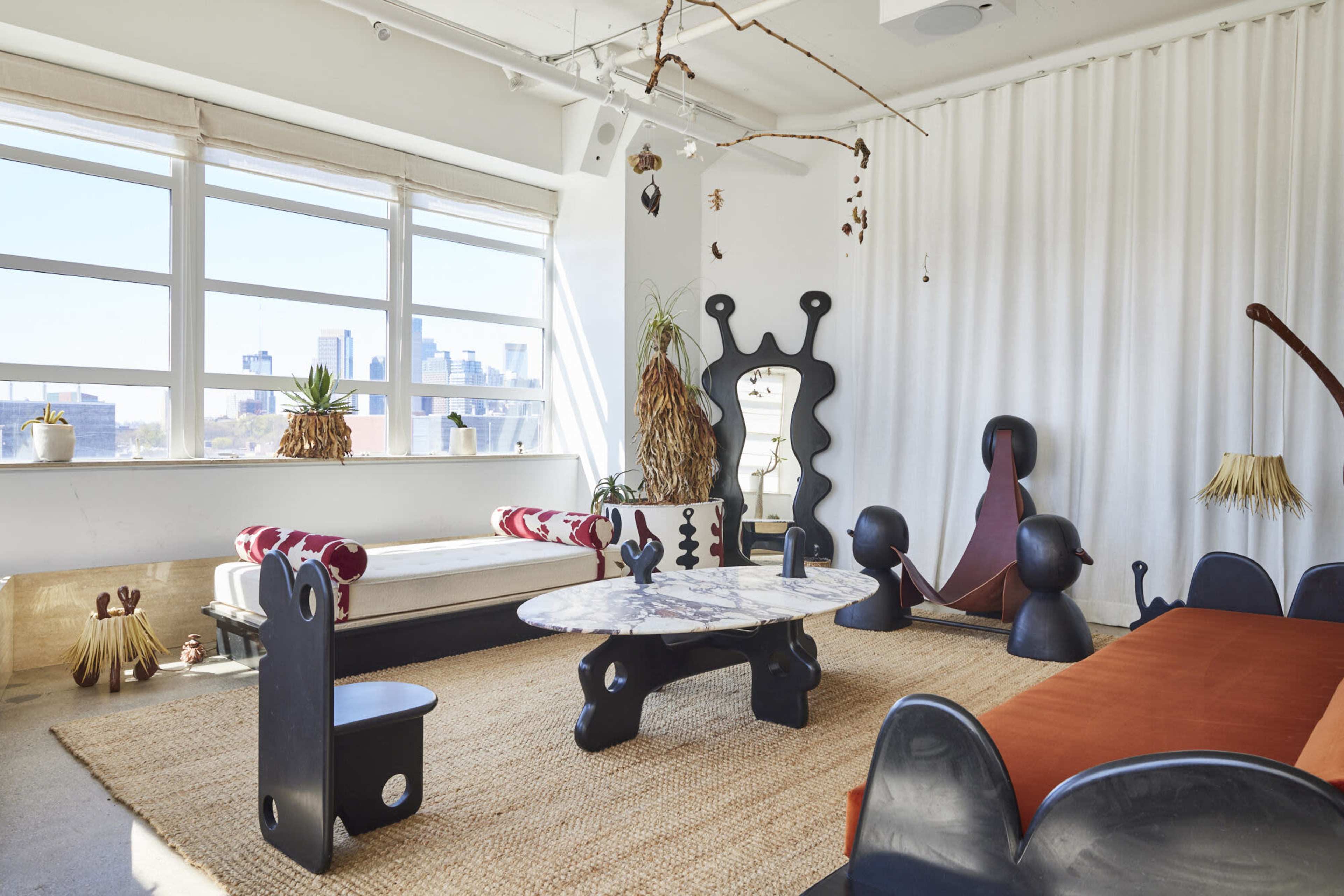 The image shows a modern living space featuring a round marble coffee table, unique sculptural furniture, and large windows that provide natural light and a view of a city skyline.