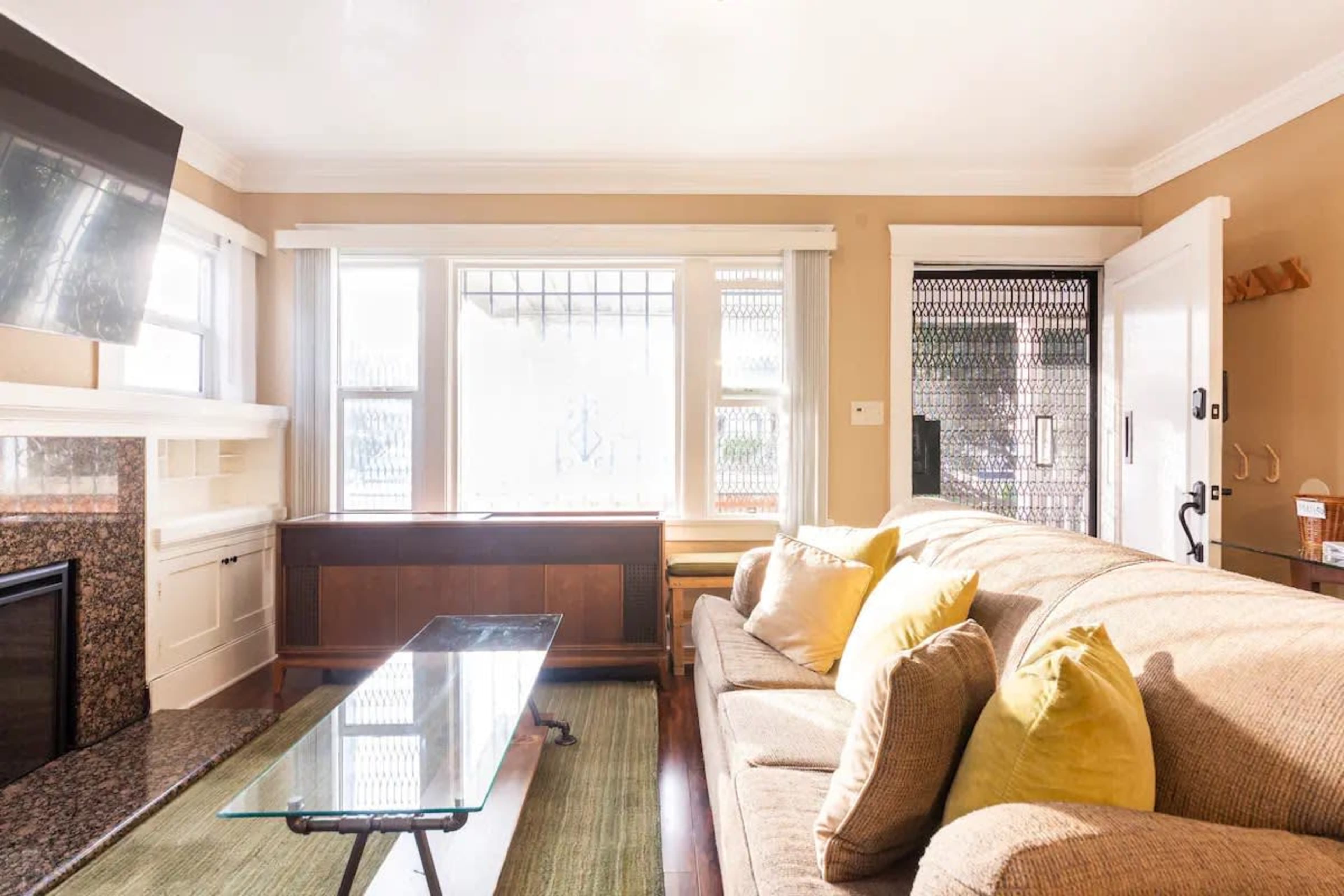 A living room features a beige couch with yellow cushions, a glass coffee table, and large windows with a view of a screened porch.
