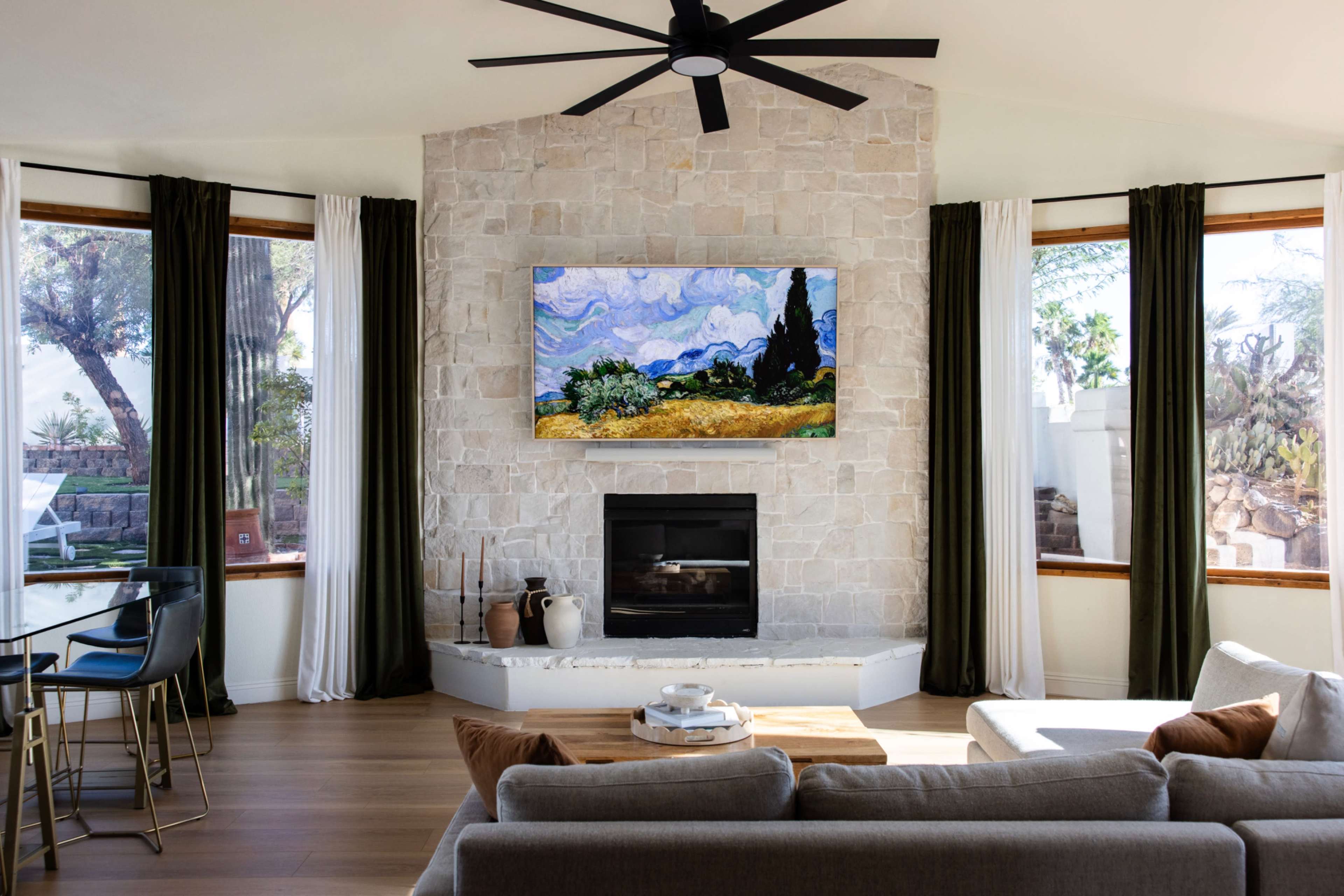 The image shows a modern living room with a stone fireplace, large windows, and a mounted artwork above the mantel.