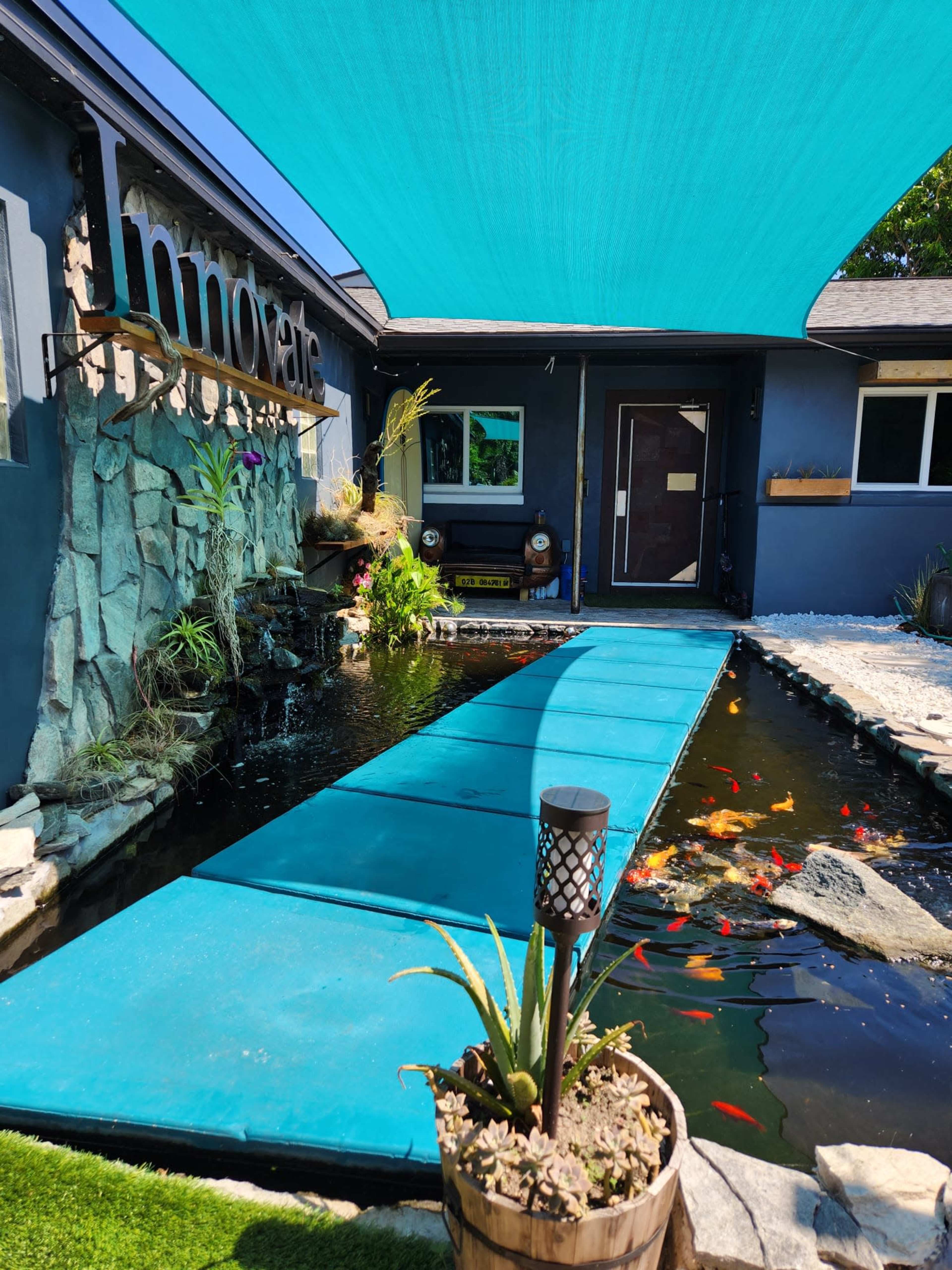 A path of blue mats leading over a pond filled with koi fish, adjacent to a modern building with a stone wall and a sign that reads "Innovate."