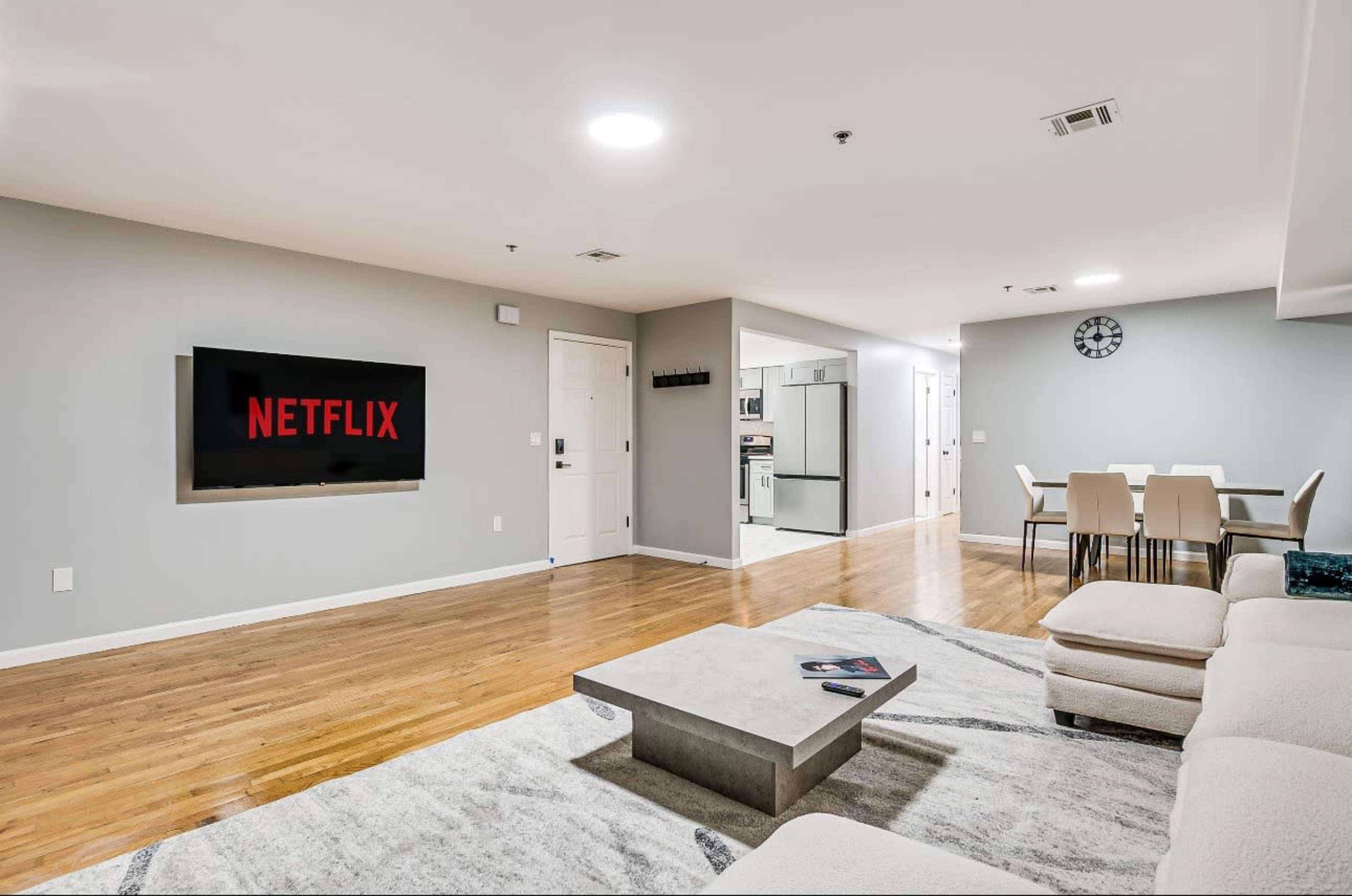 The image shows a modern living room with a large flat-screen TV displaying the Netflix logo, a light-colored sectional sofa, a coffee table, and a dining area with a table and chairs visible in the background.
