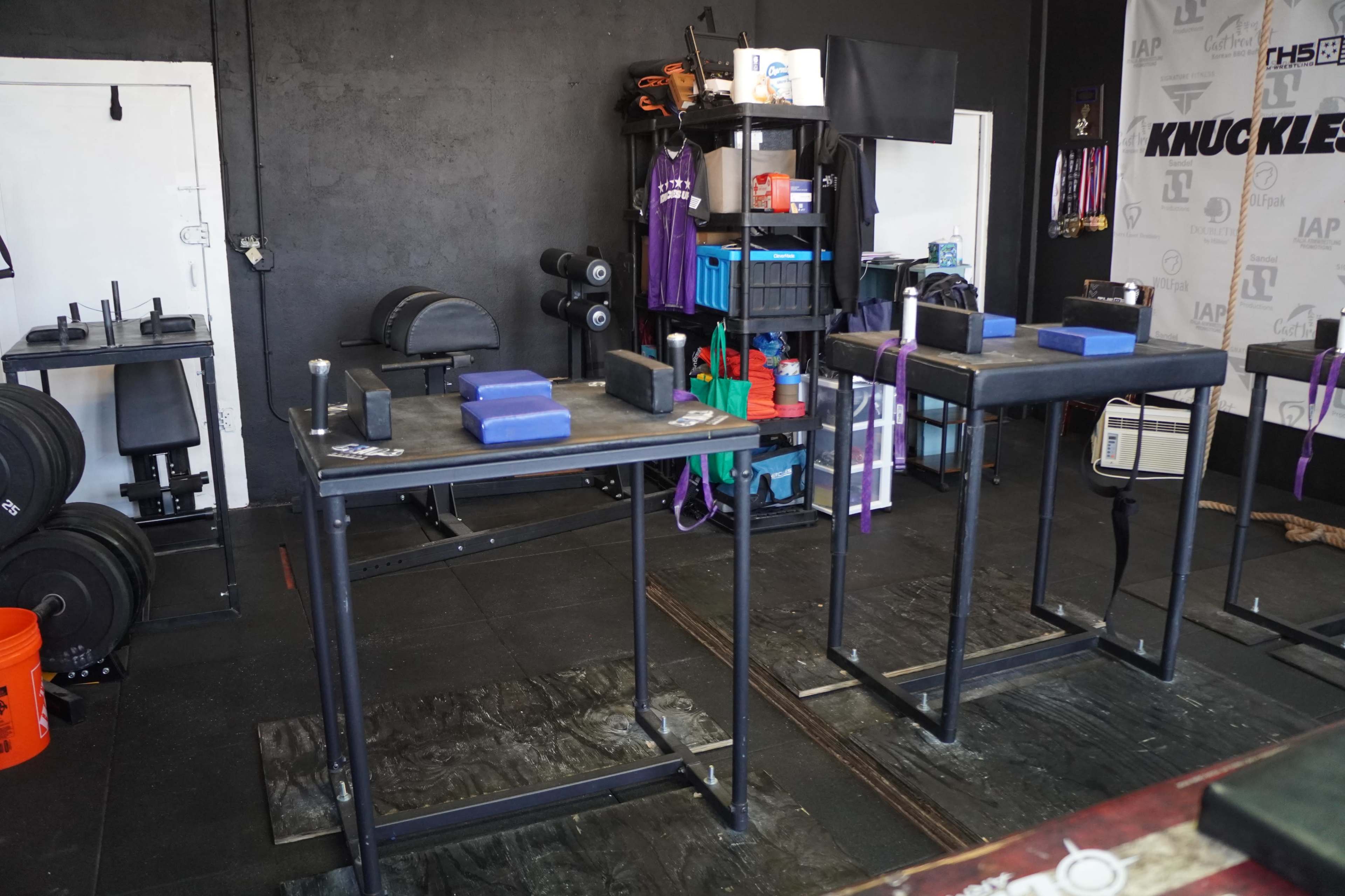 The image shows a gym interior featuring exercise equipment, including two parallel bars, mats, weights, and storage racks against a black wall.