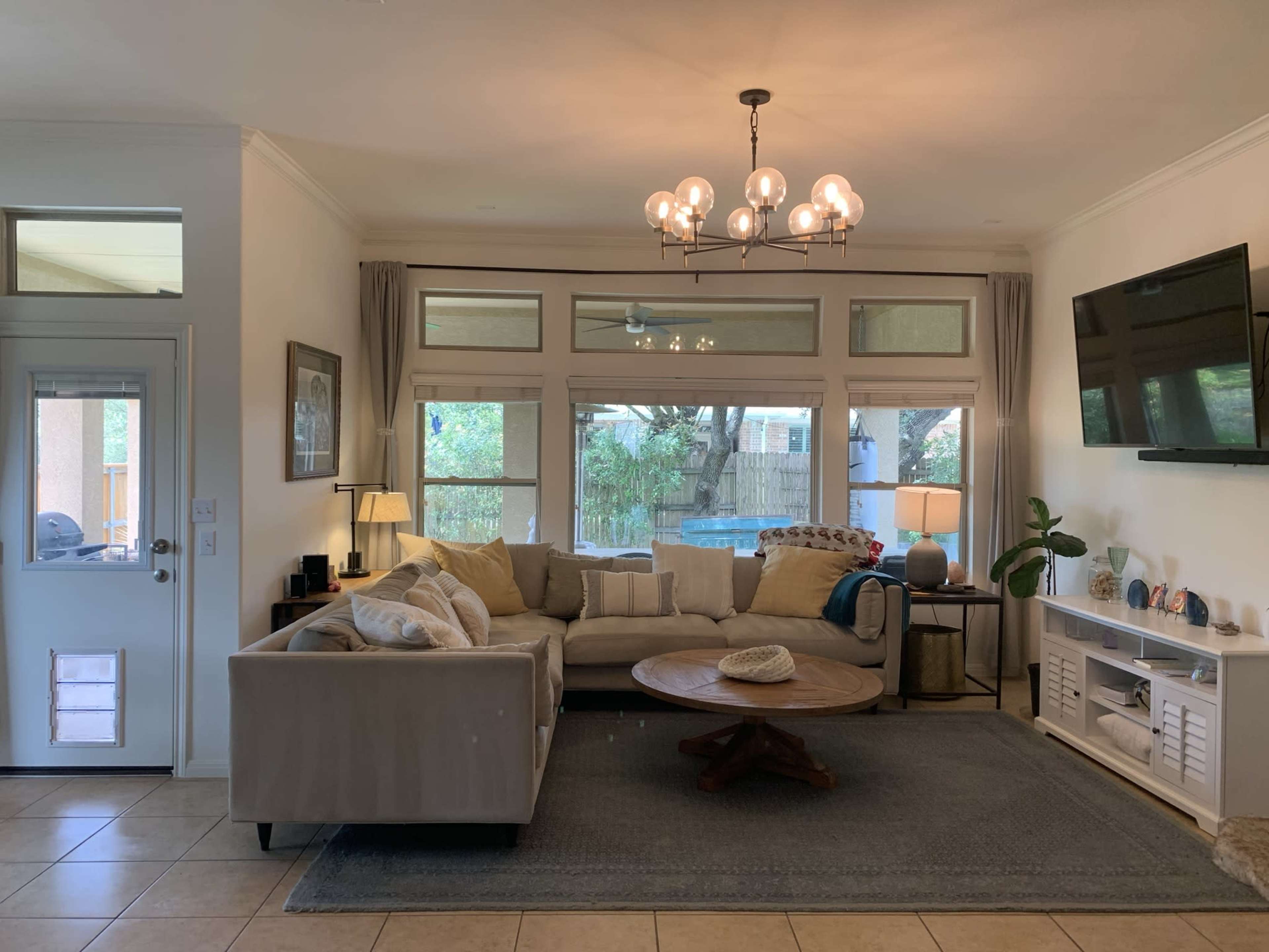 A living room features a large sectional sofa, a round coffee table, a TV mounted on the wall, and large windows providing a view of the outdoors.