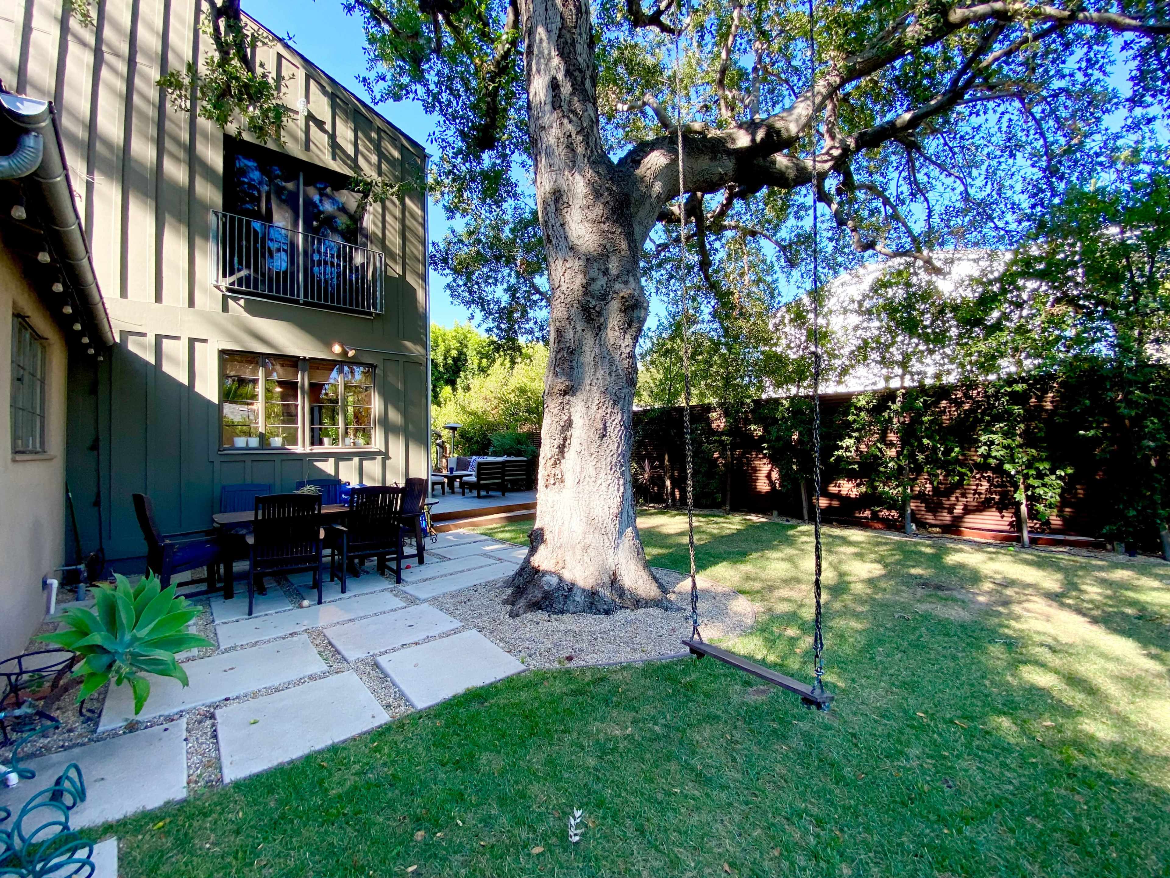 A large tree with a swing stands in a backyard adjacent to a house, featuring a stone pathway and a patio set.