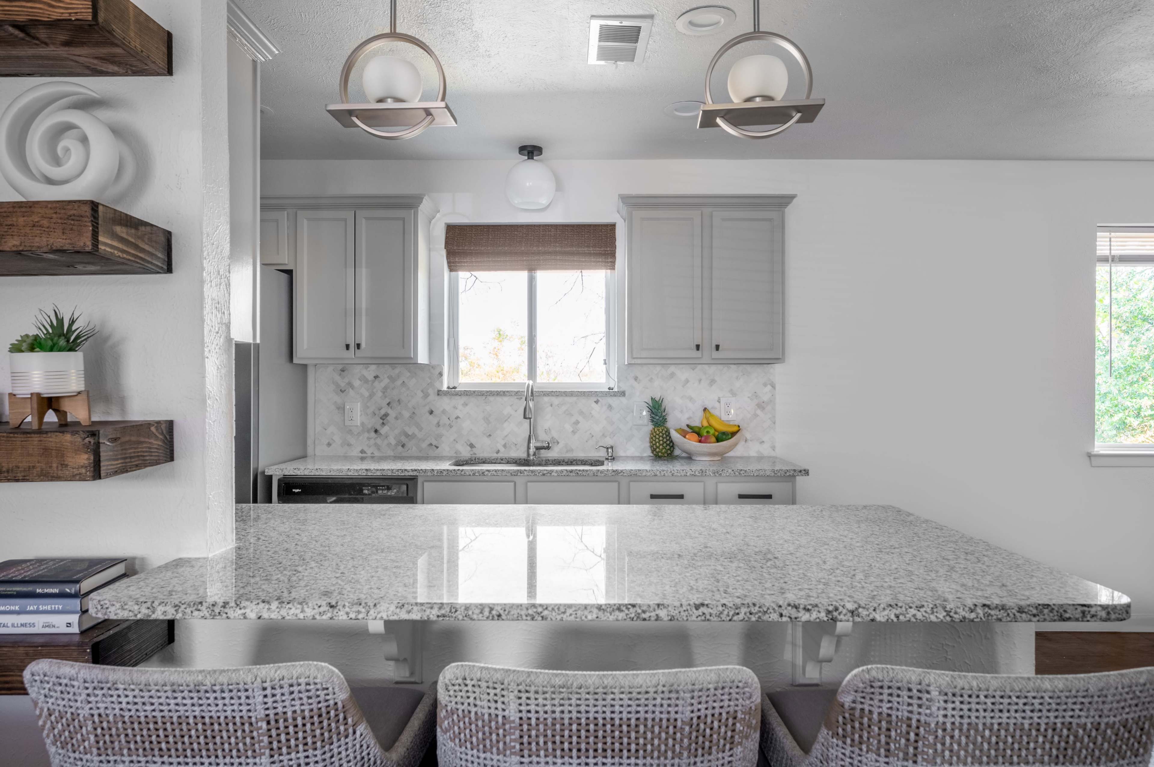 A modern kitchen features gray cabinetry, a granite countertop with bar seating, and a window overlooking a garden.