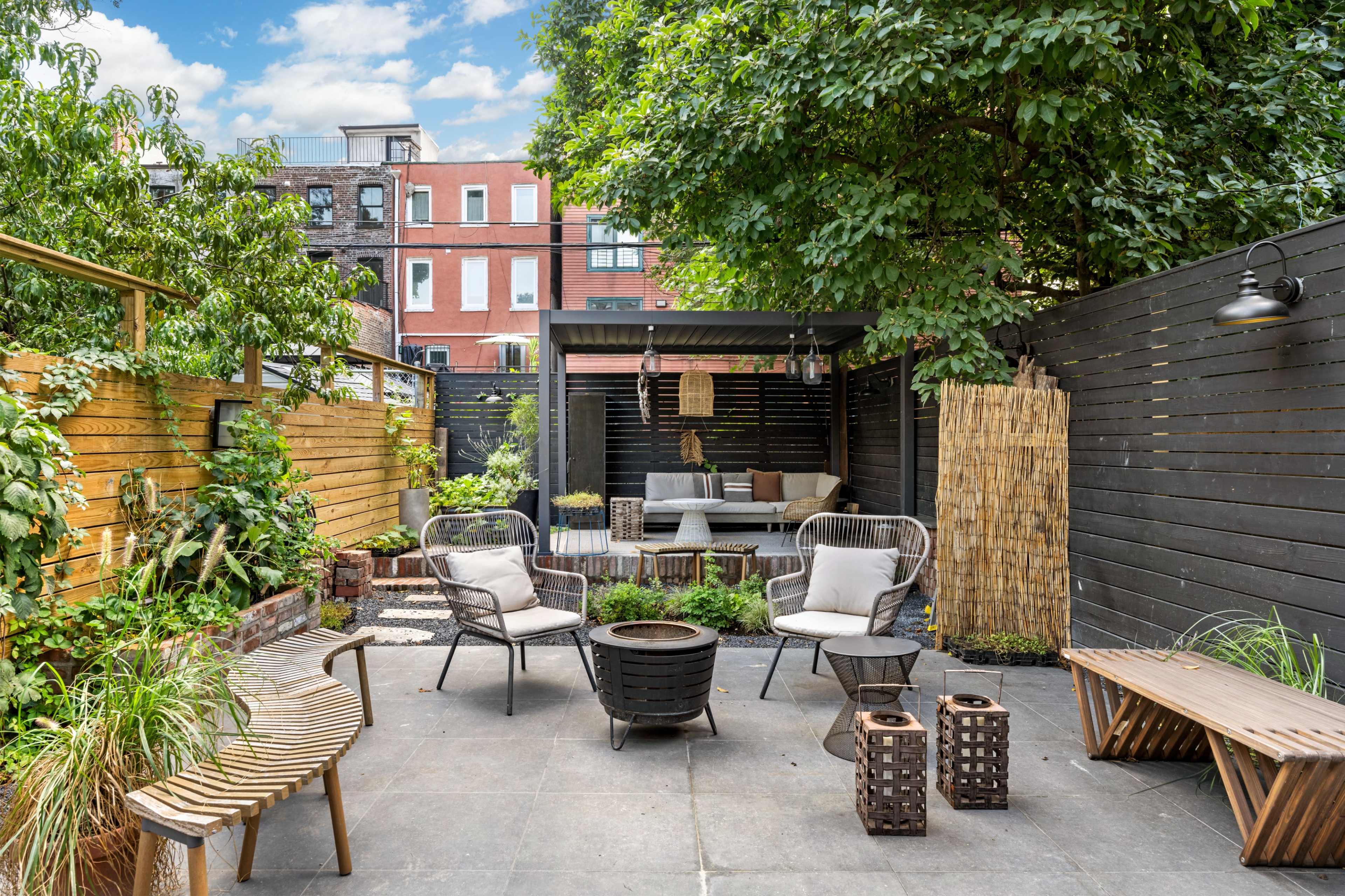 A small urban patio features seating areas, a fire pit, and greenery surrounded by wooden and black privacy fences.