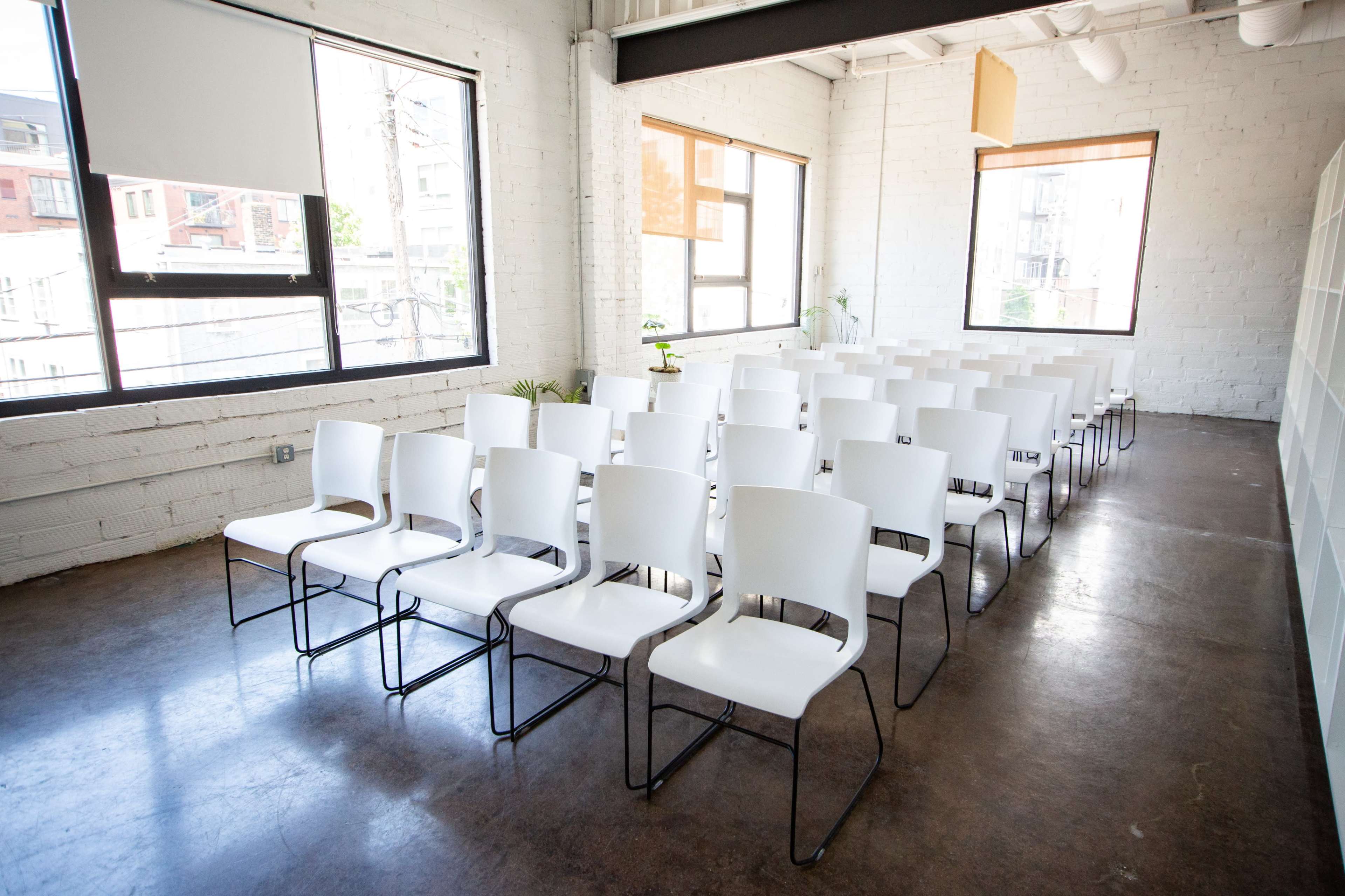 The image shows a minimalist room with rows of white chairs set up for an event, illuminated by natural light from large windows.