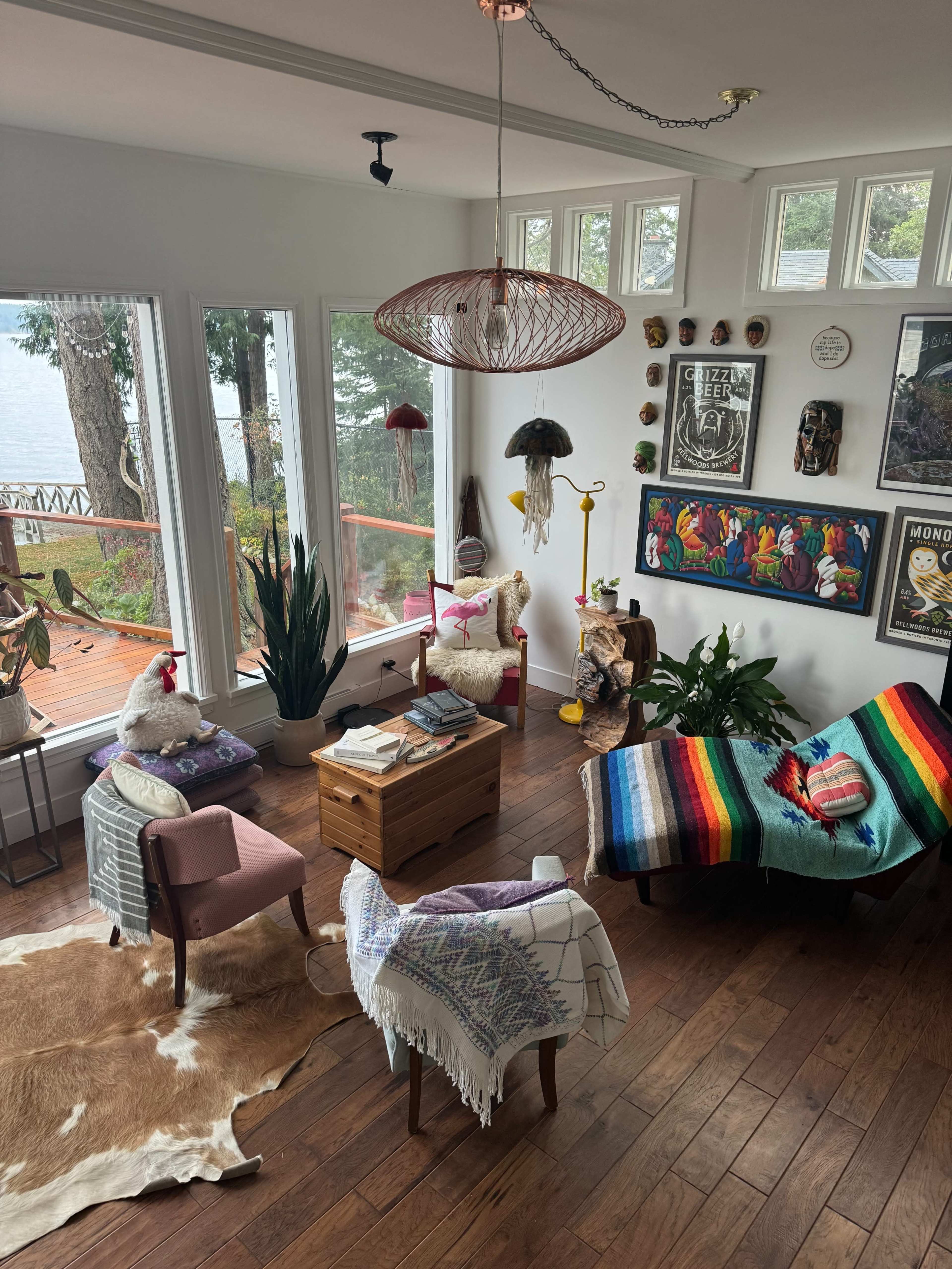 A cozy living room with large windows overlooking a lake, featuring a mix of eclectic furniture, plants, and colorful decorations.