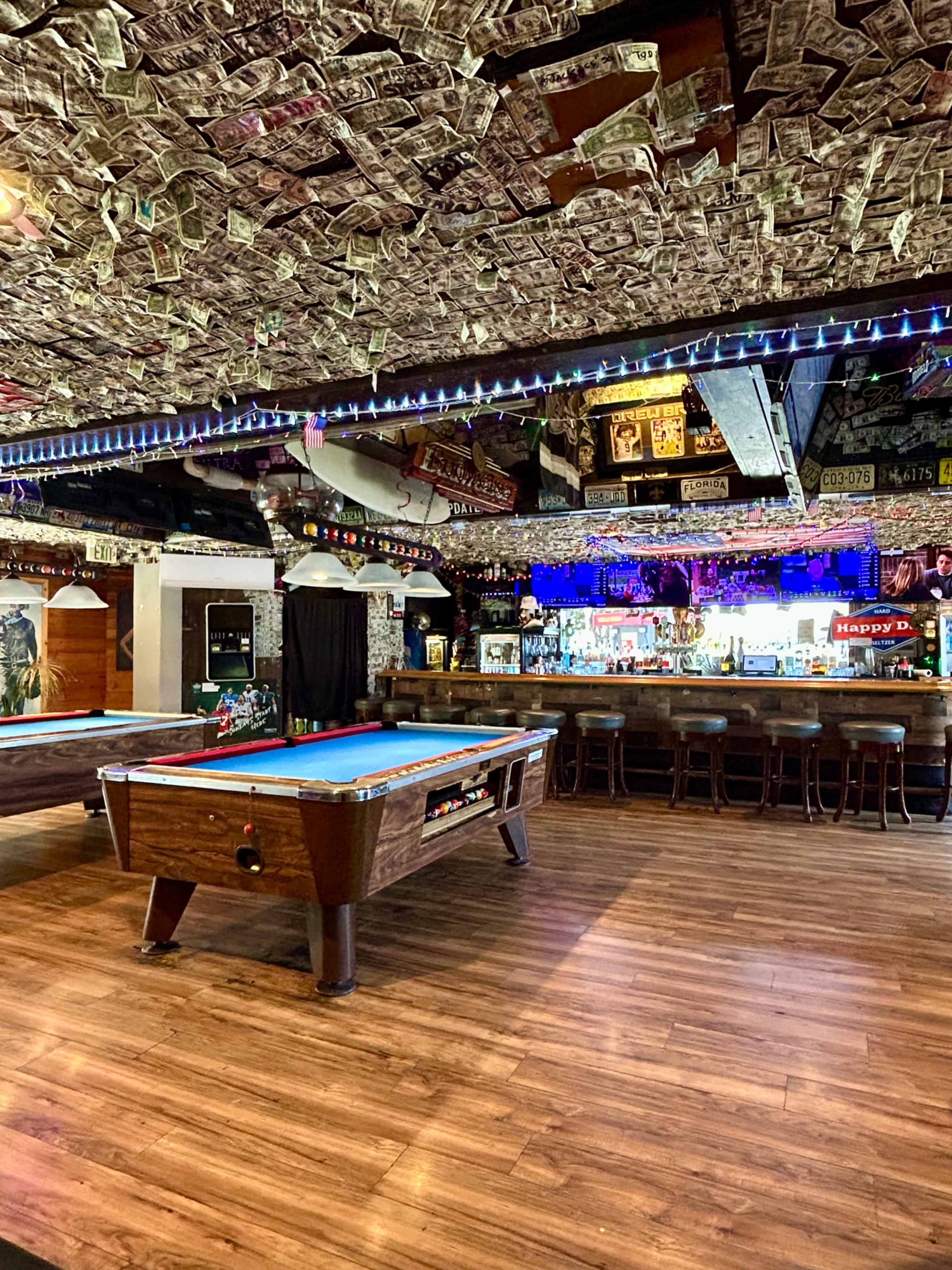 The image shows a bar with a pool table in the foreground, colorful lights, and a ceiling covered in dollar bills.