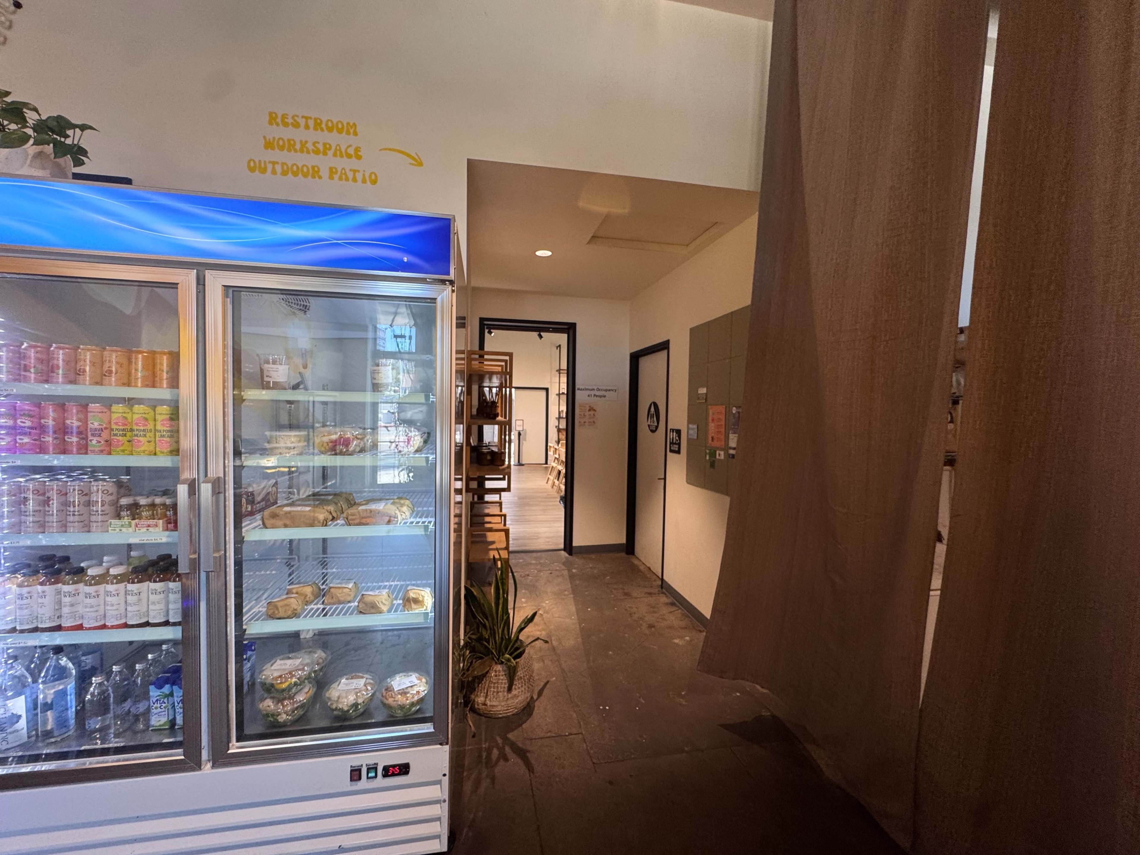 The image shows a hallway with a refrigerator filled with drinks and snacks on the left, leading to a doorway labeled for restrooms and other facilities.