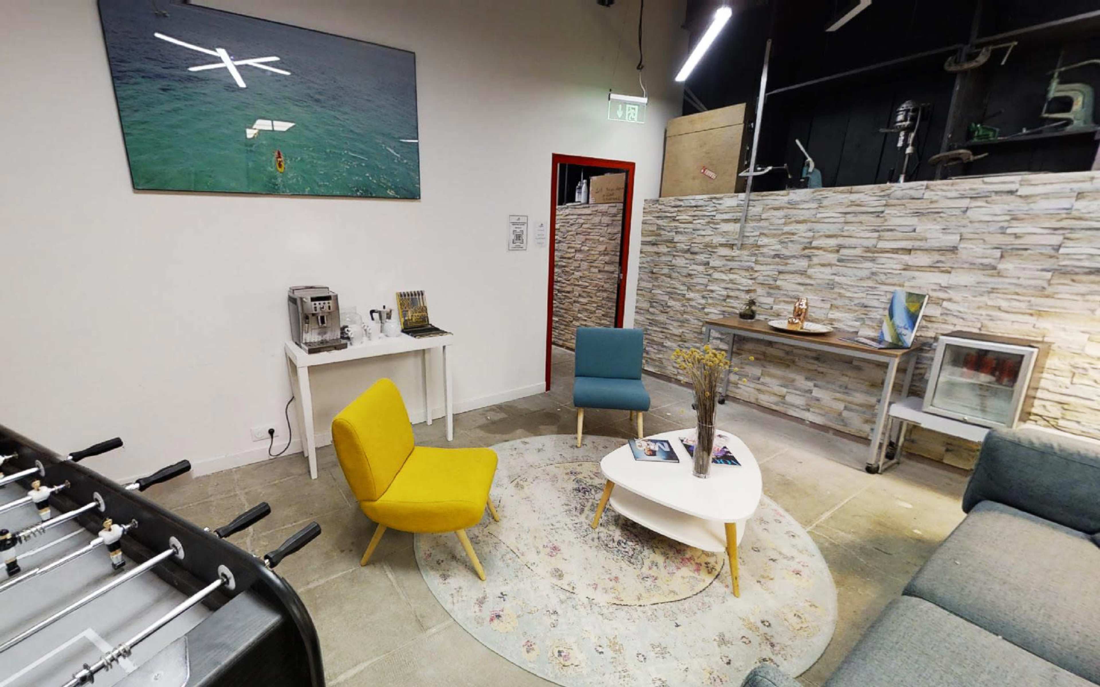 The image shows a modern lounge area featuring a yellow and a blue chair, a white coffee table on a patterned rug, and a gray sofa against a textured stone wall.
