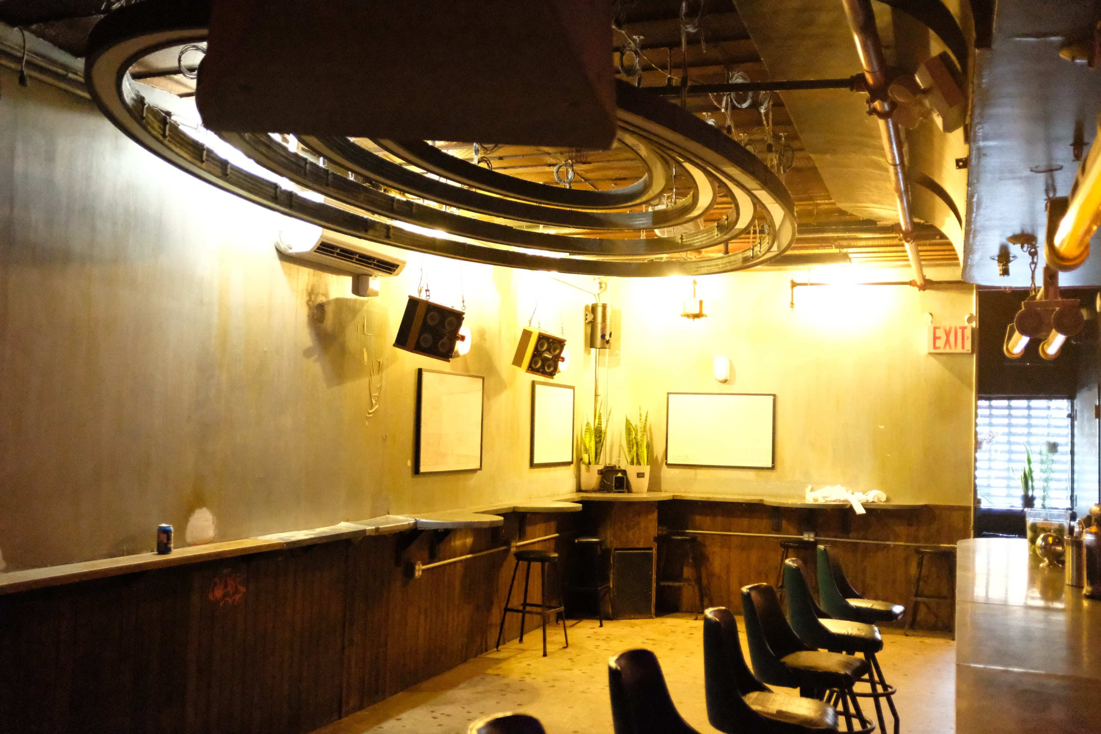The image shows an indoor space with a bar area, featuring hanging speakers, a circular light fixture, and a few chairs against the wall.