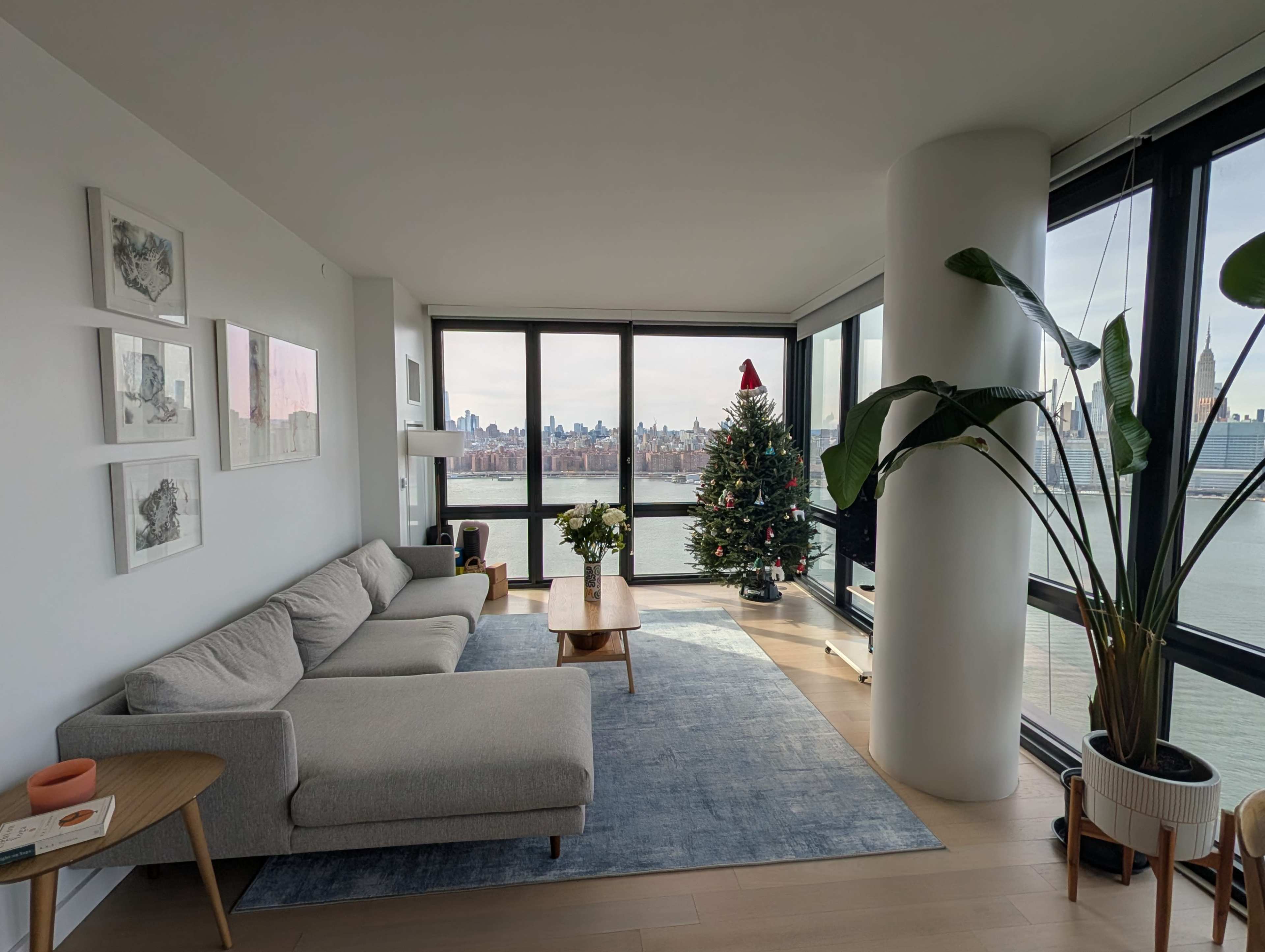 A spacious living room features a gray sofa, a wooden coffee table, a large Christmas tree adorned with decorations, and panoramic windows showcasing a city skyline.