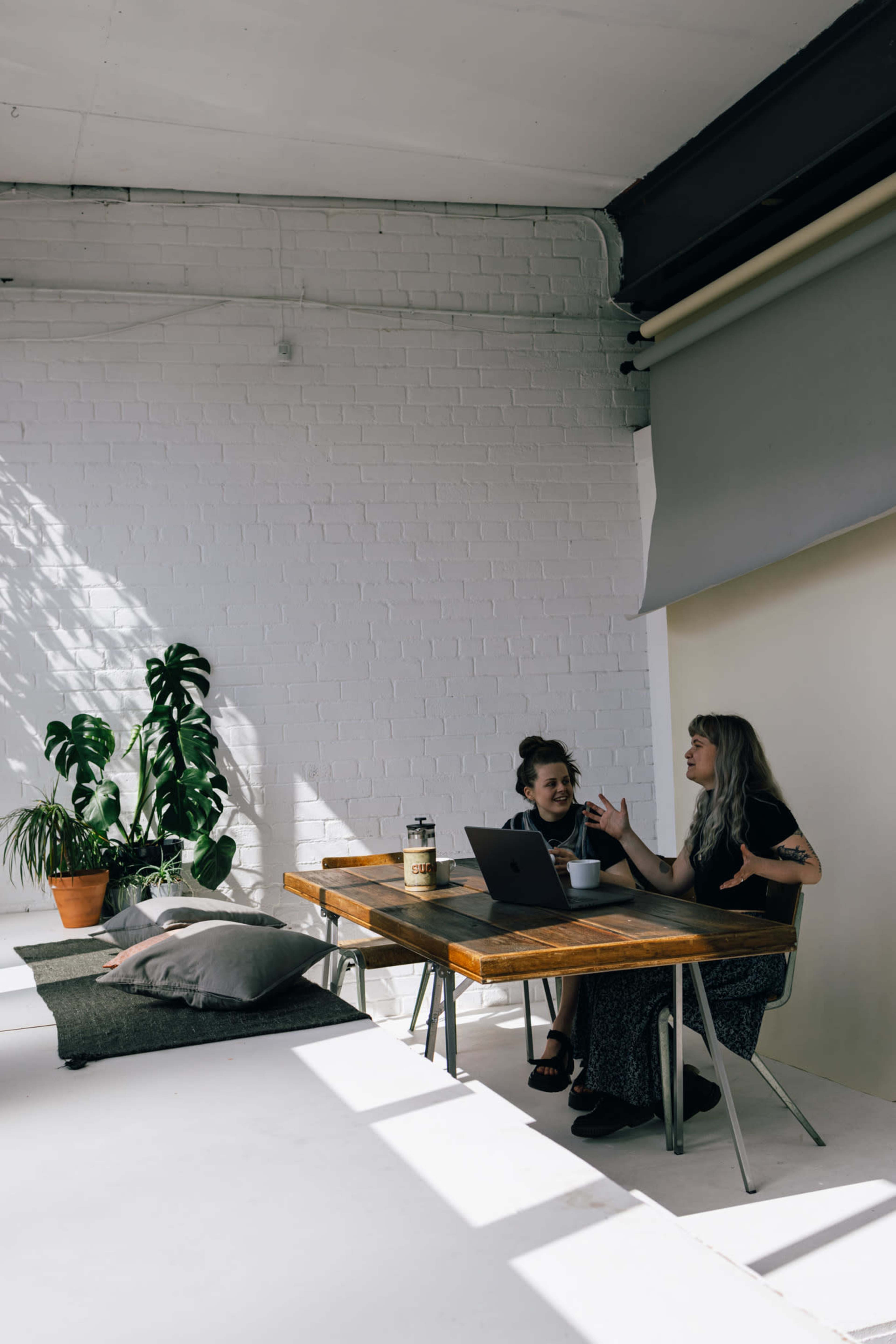 Two women engage in conversation at a wooden table in a bright, minimalistic workspace, with a large plant nearby and soft cushions on the floor.