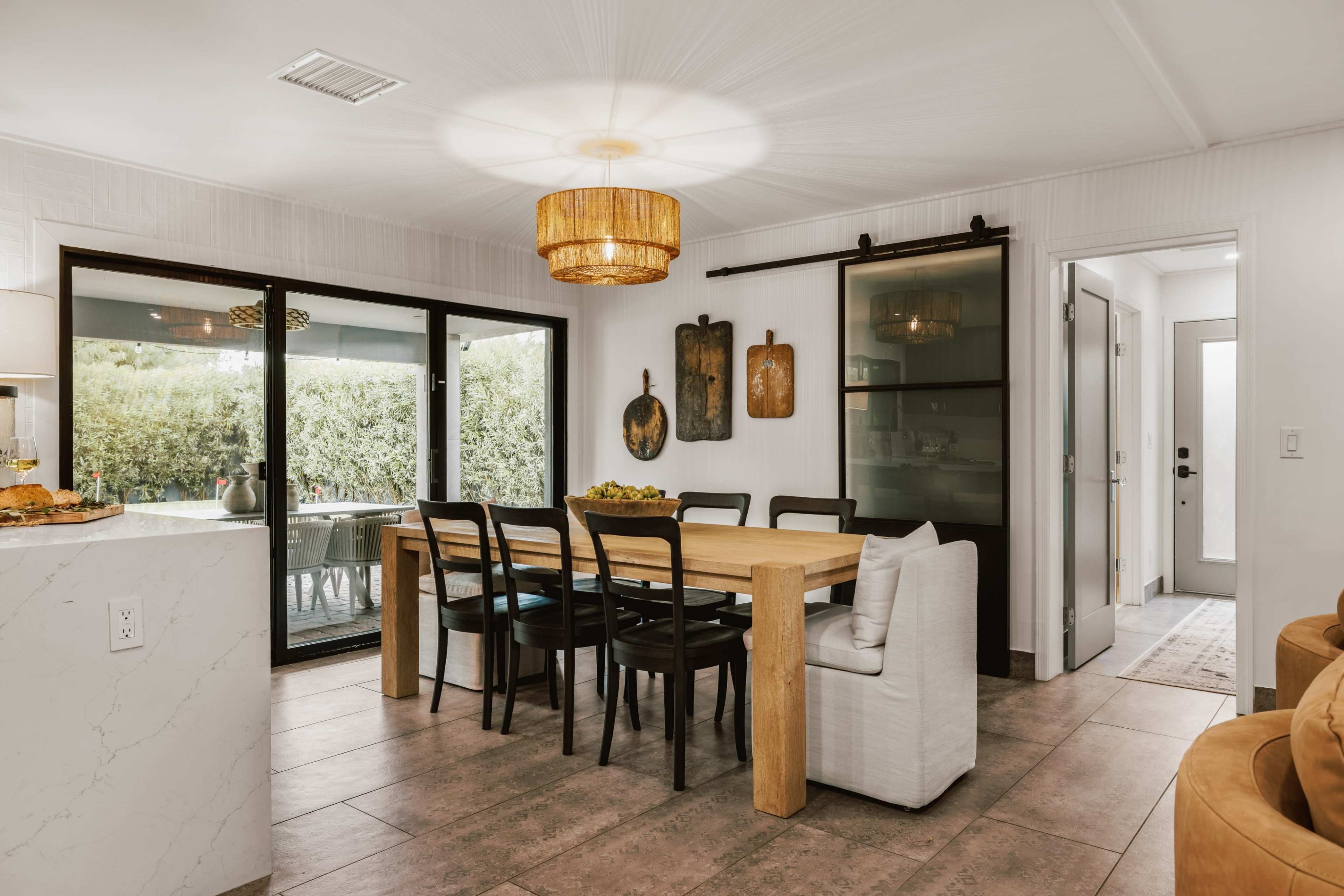 The image shows a modern dining area with a wooden table surrounded by black chairs, a ceiling light fixture, and large glass doors leading to an outdoor space.