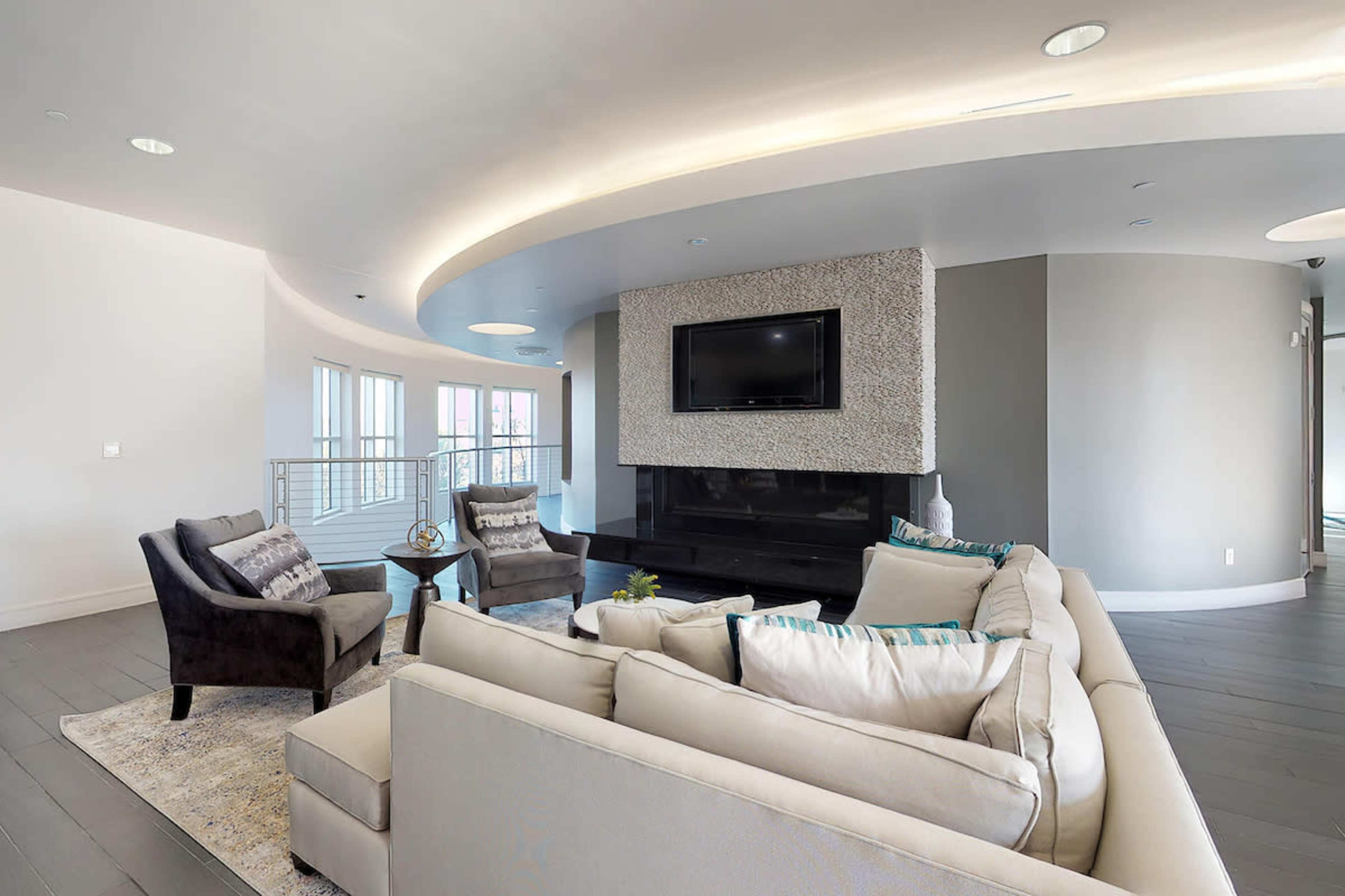 A modern living room features a curved ceiling, a minimalist fireplace, a mounted TV, and contemporary furniture arranged around a central area rug.
