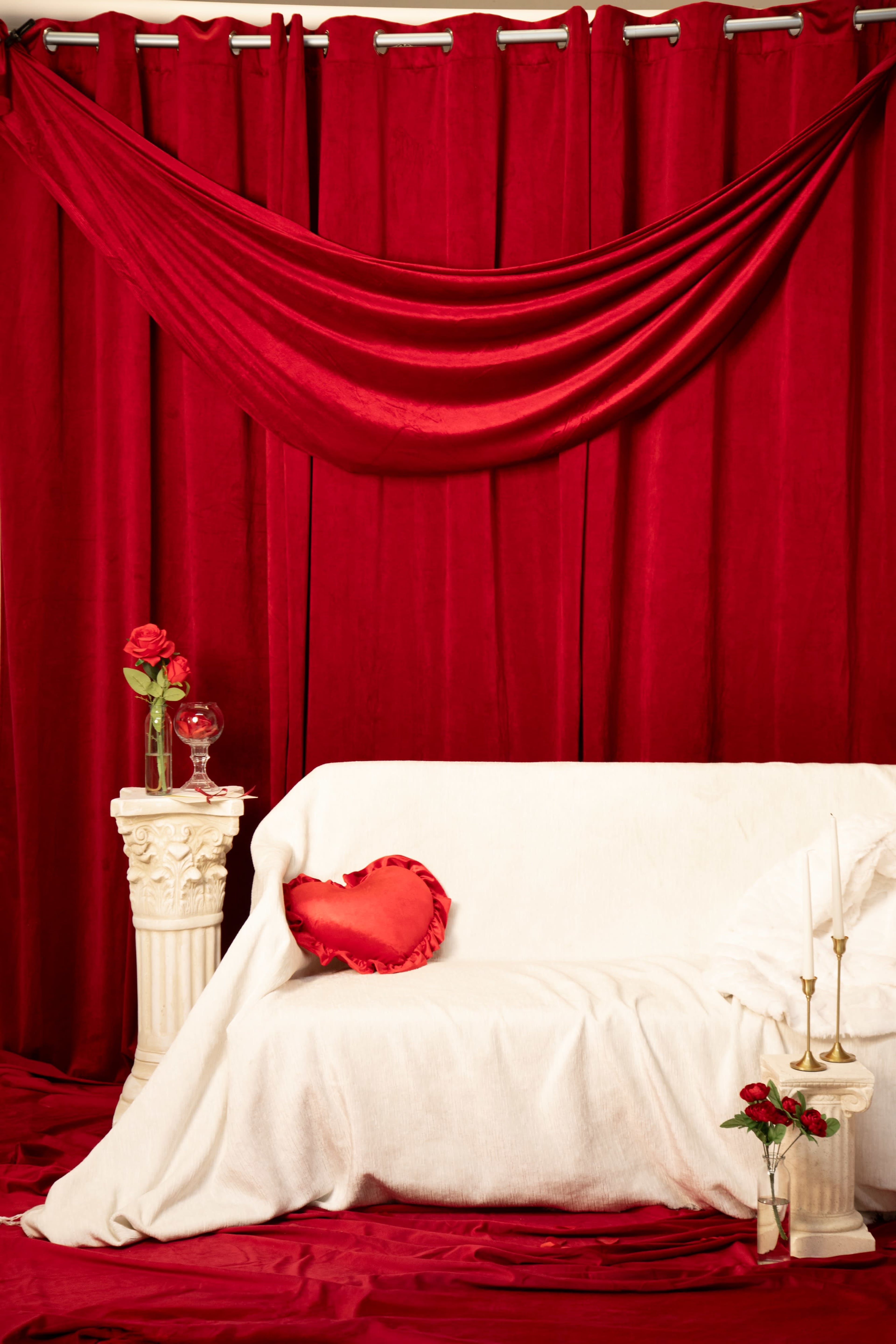 The scene features a white sofa draped in a white cloth, placed against a backdrop of red curtains, with decorative elements including roses, a glass, and candles on a pedestal.