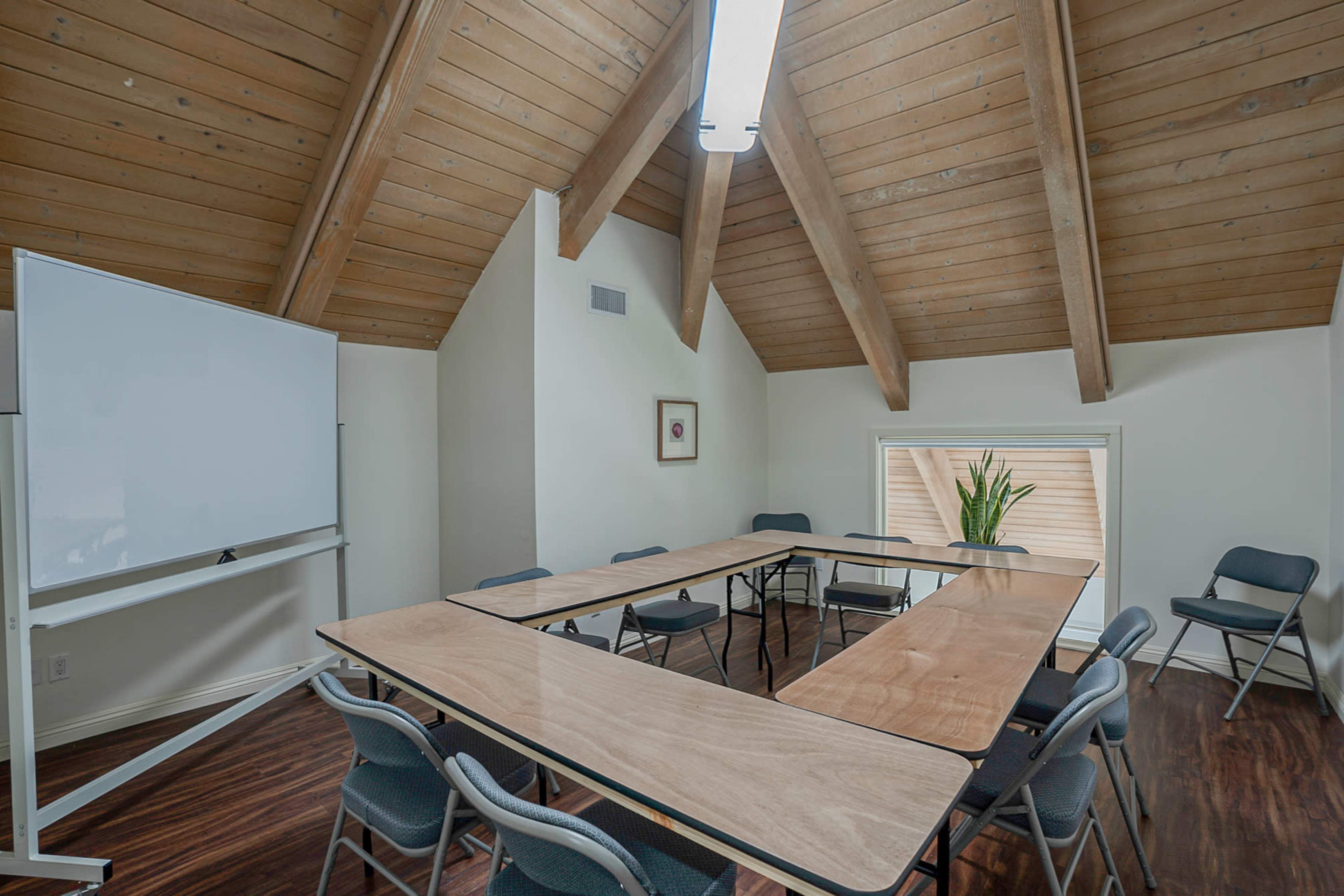A conference room features a large wooden table arranged in a U-shape, surrounded by gray chairs, with exposed wooden beams overhead and a whiteboard to one side.