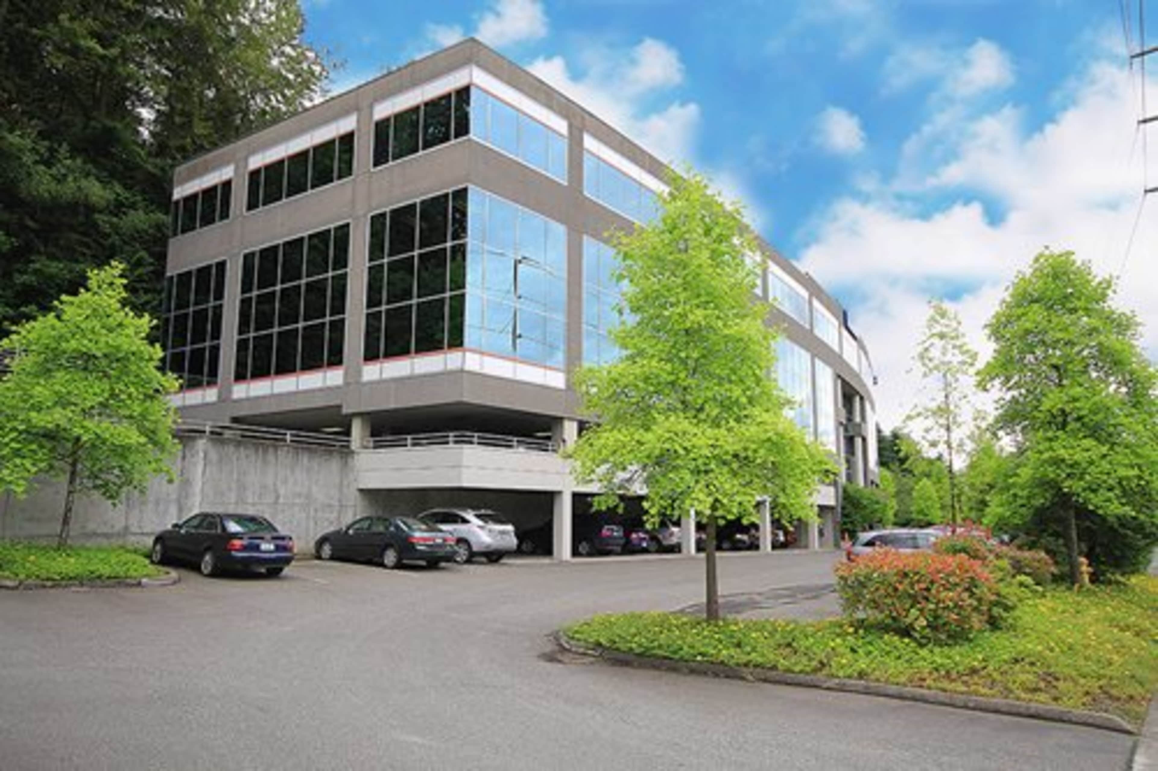 A modern office building with large glass windows and an adjacent parking area surrounded by greenery.