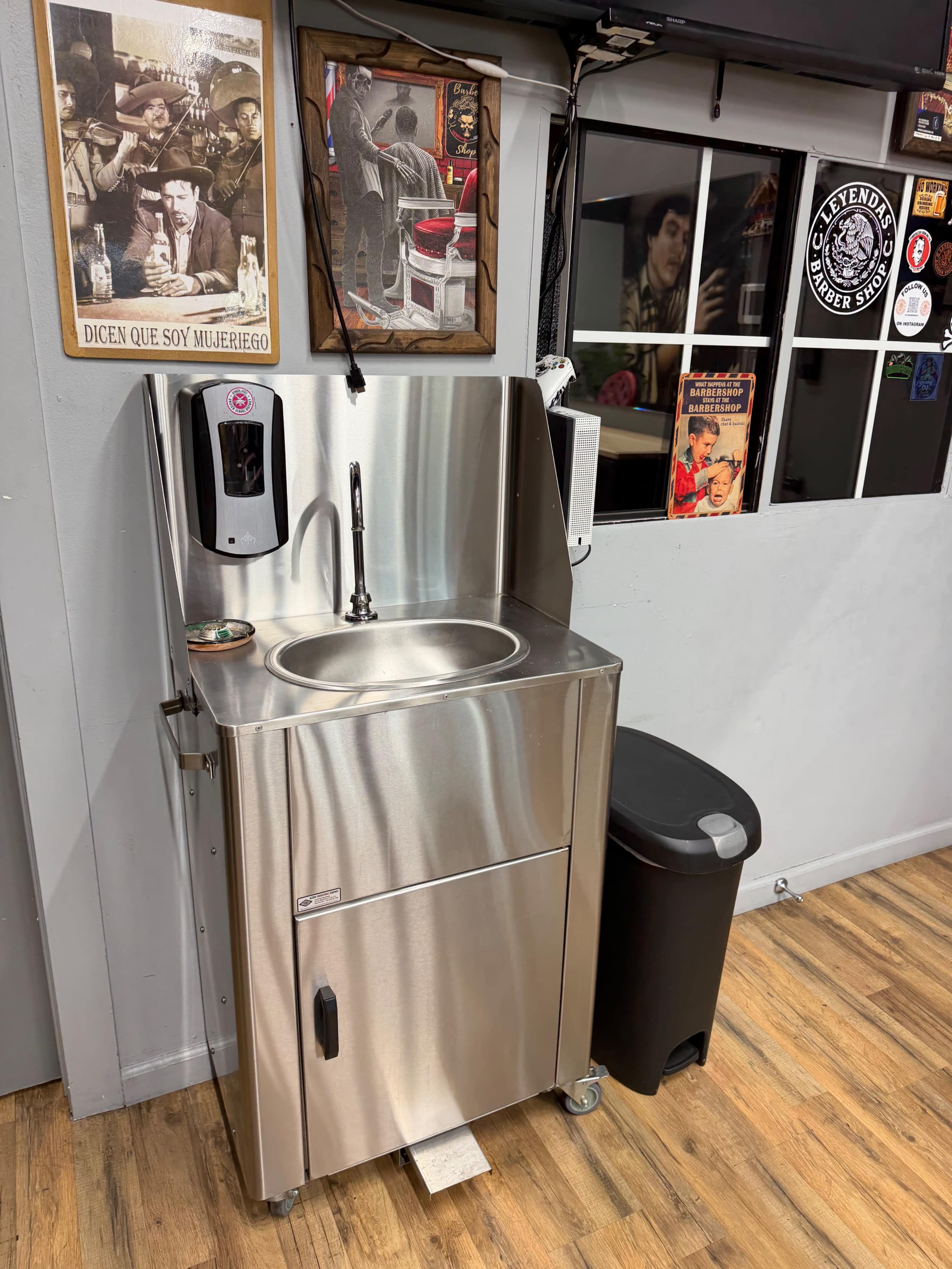 The image shows a compact stainless steel sink station with a hand sanitizer dispenser, located near a wall adorned with various posters and a trash can nearby.
