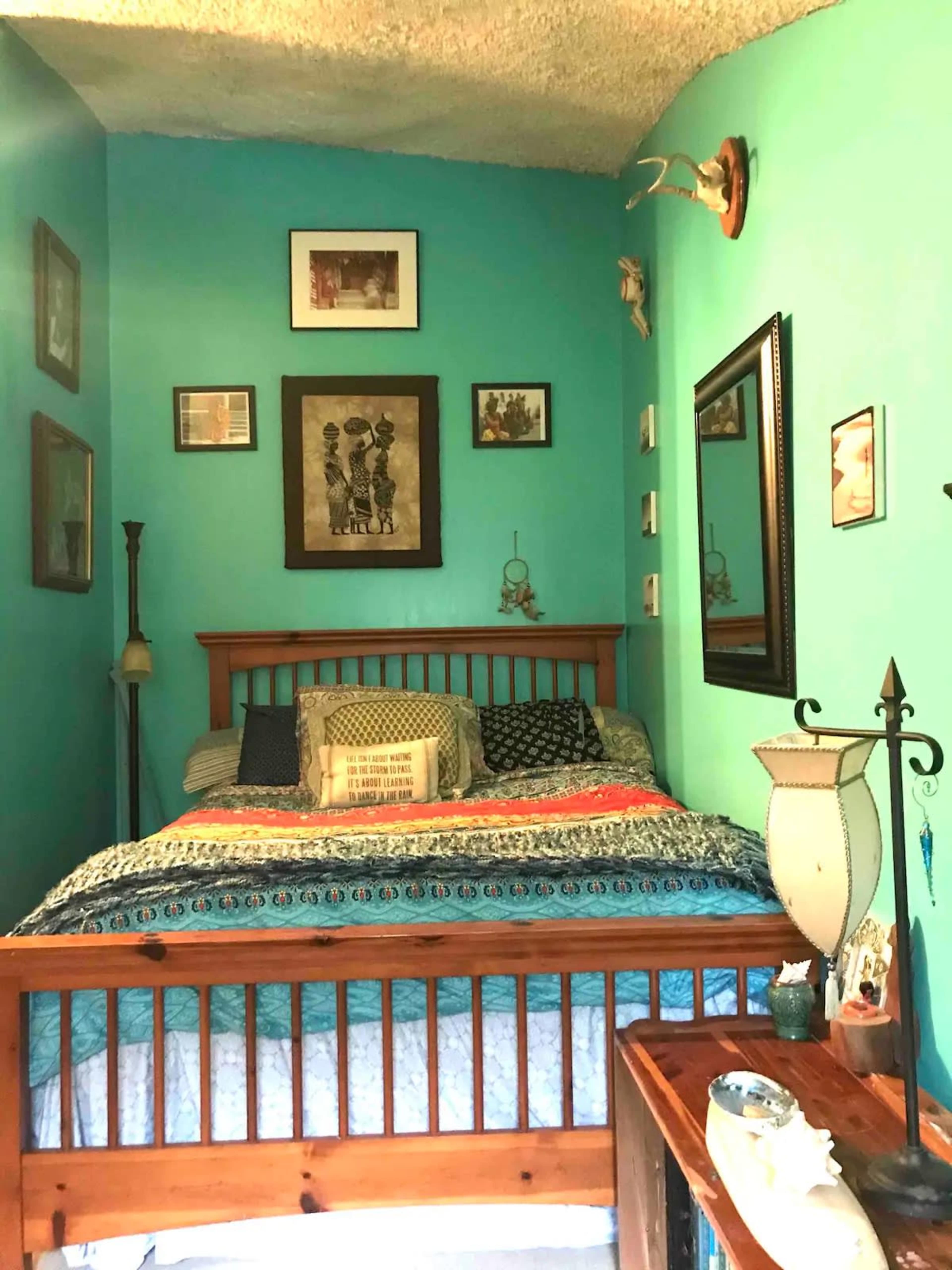 The image shows a small bedroom with a wooden bed, turquoise walls, and various framed pictures and decorative items arranged around the room.