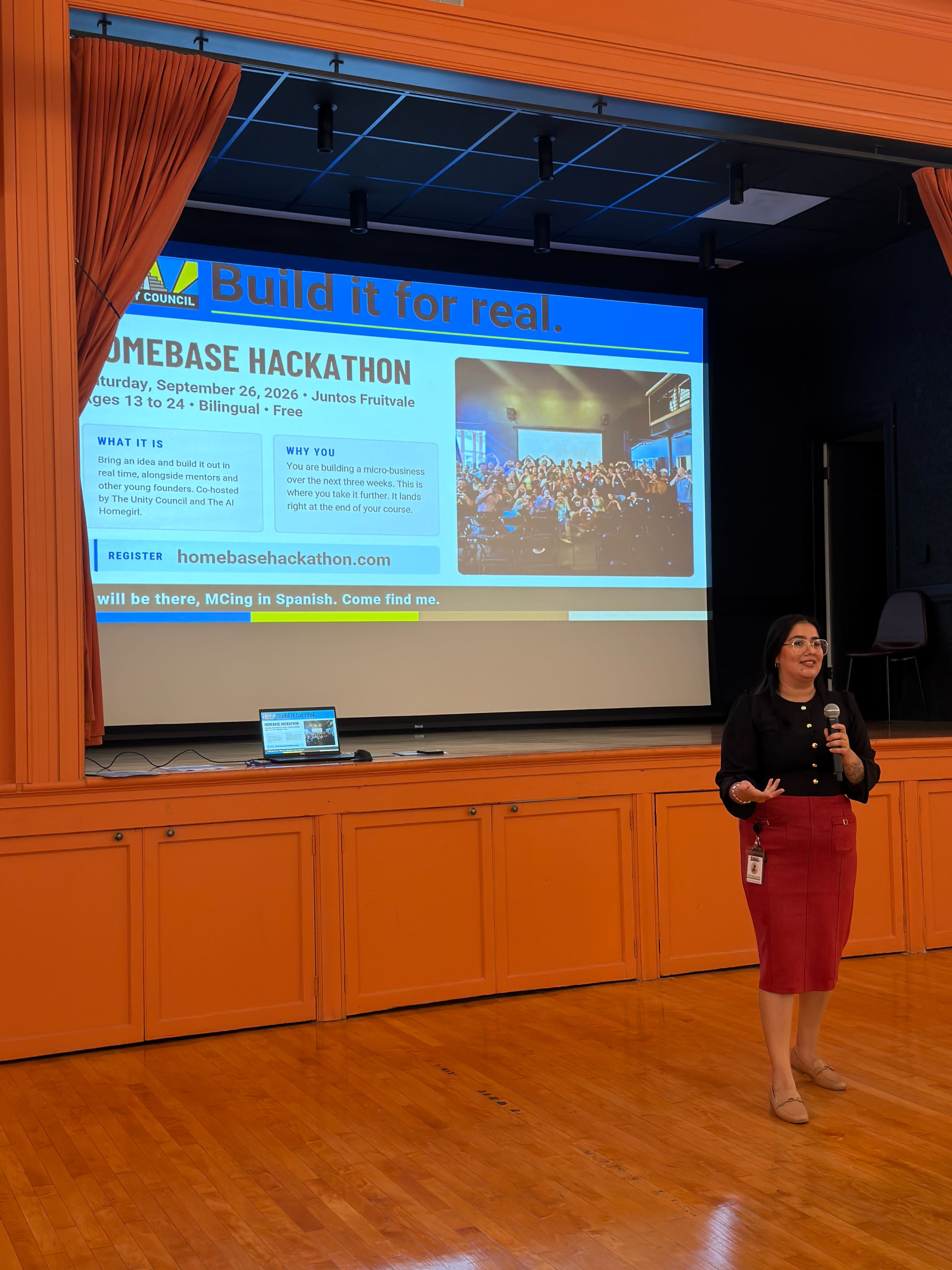 A woman stands on stage in a brightly lit auditorium, presenting information about the Homebase Hackathon on a large screen behind her.