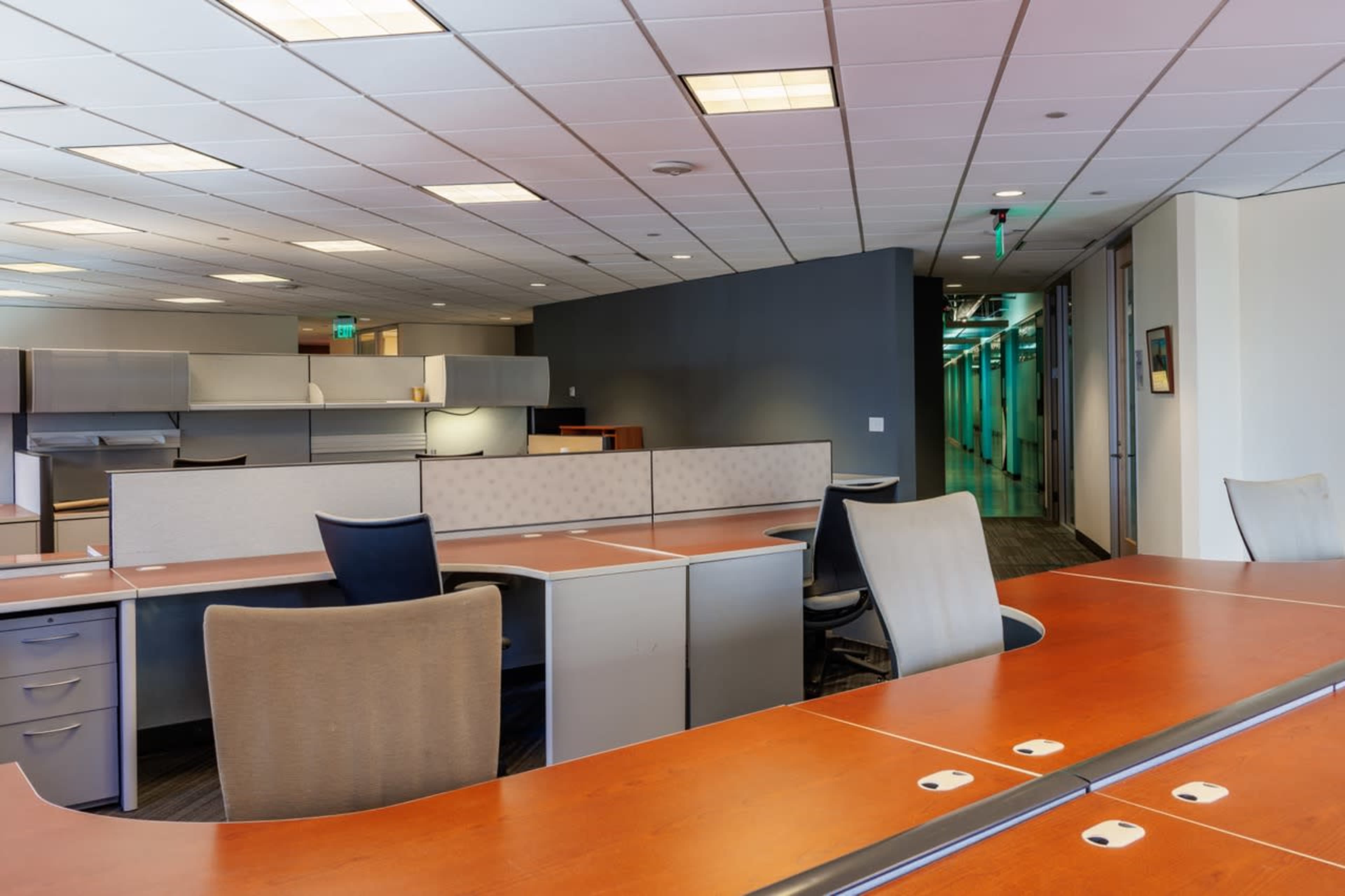 An office space features several empty workstations with desks and ergonomic chairs, along with gray partition walls.