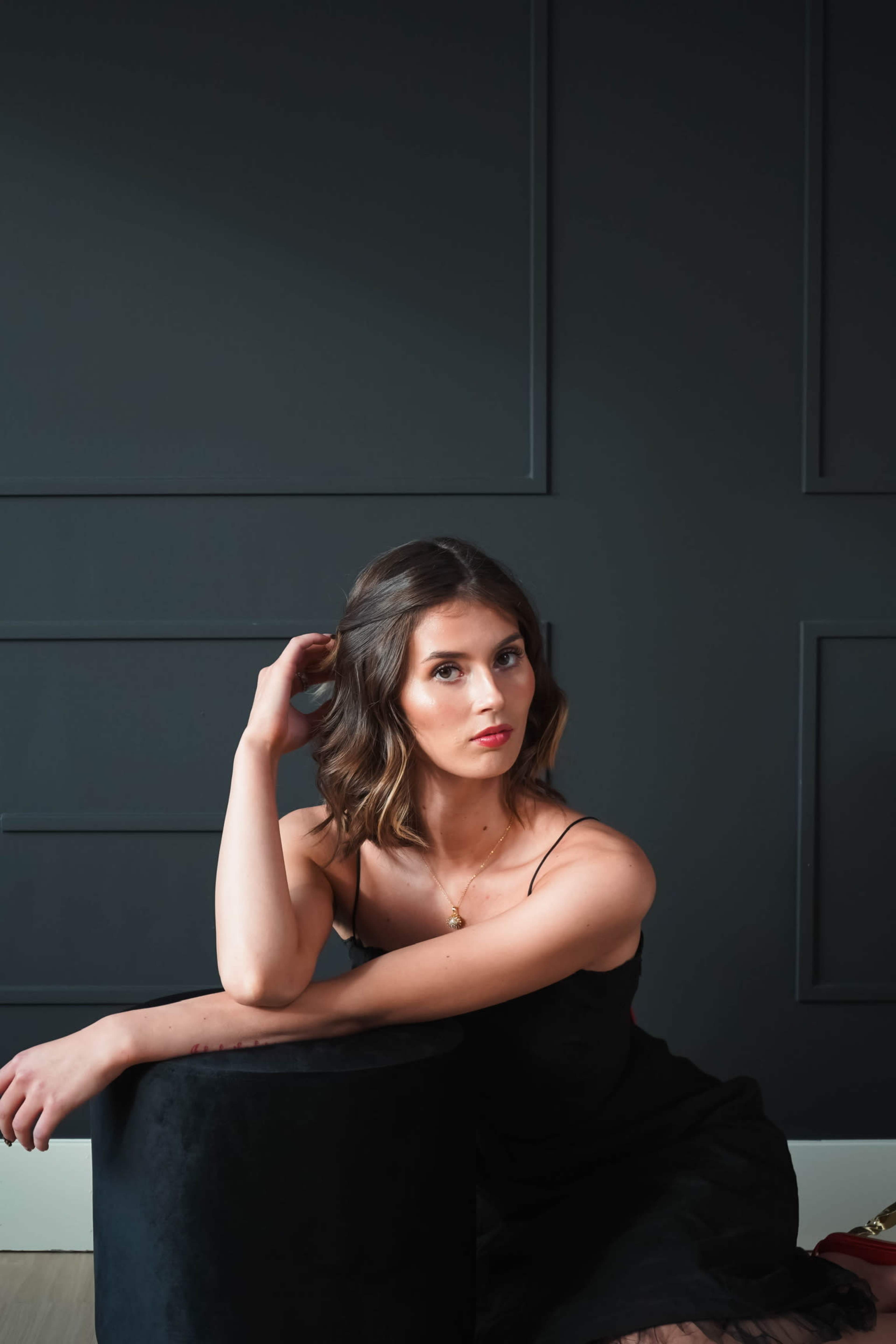 A woman with shoulder-length hair is sitting on a black cylindrical stool against a dark wall, resting her elbow on her knee.