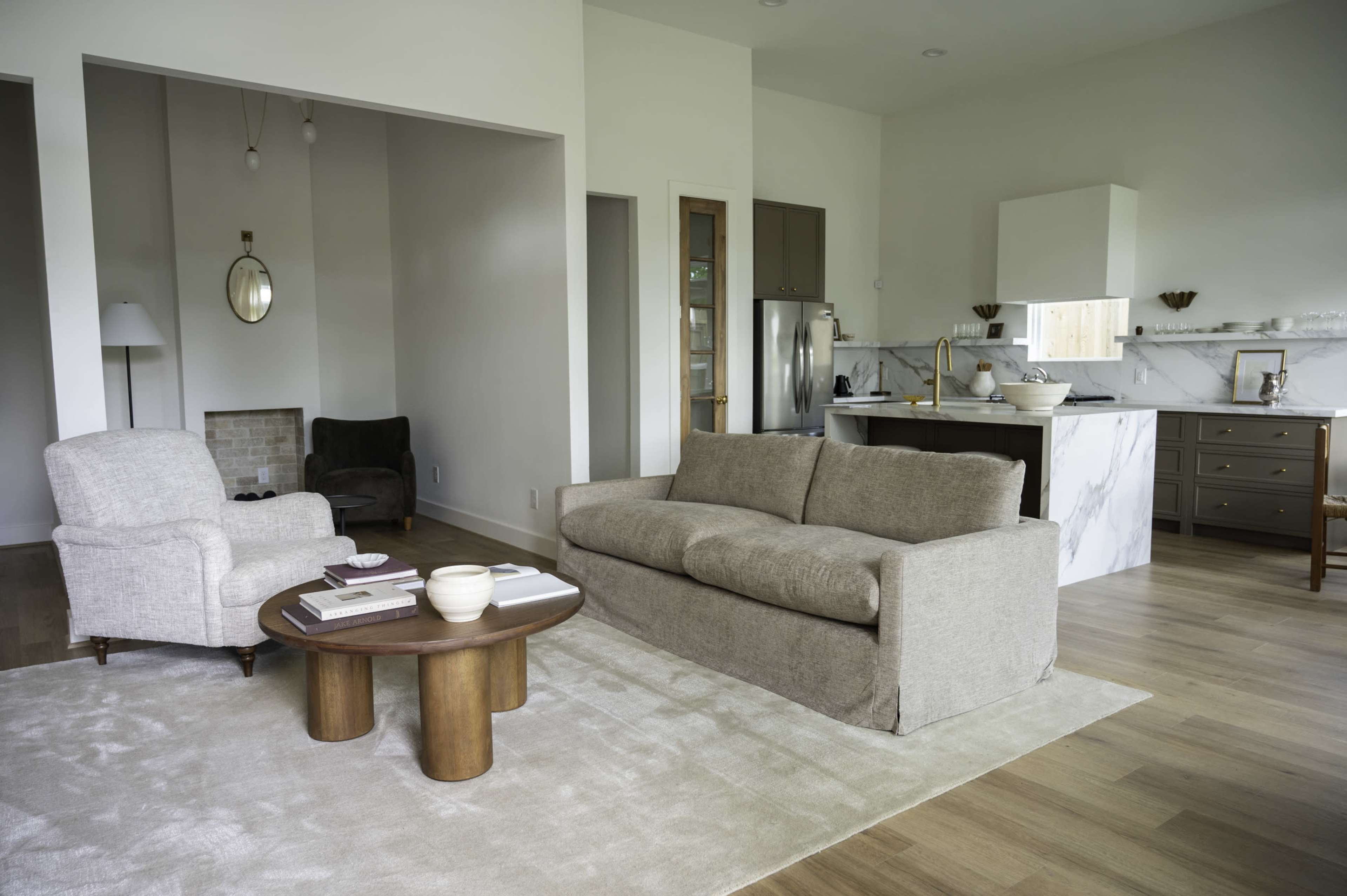 The image shows a modern living room with a beige sofa, two armchairs, a round wooden coffee table, and a kitchen area visible in the background.