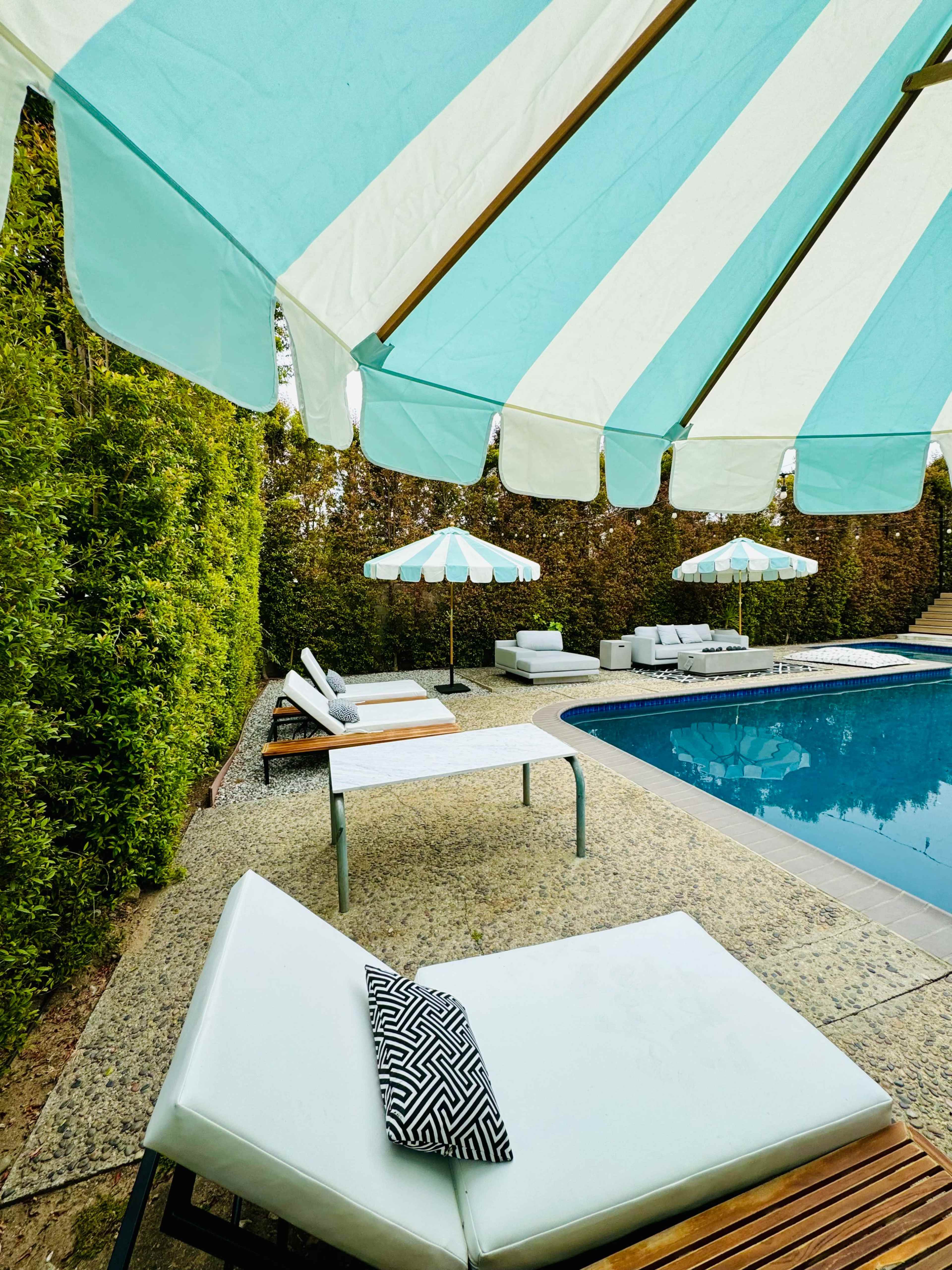 A pool area features striped umbrellas, lounge chairs, and a clear blue water surface surrounded by greenery.