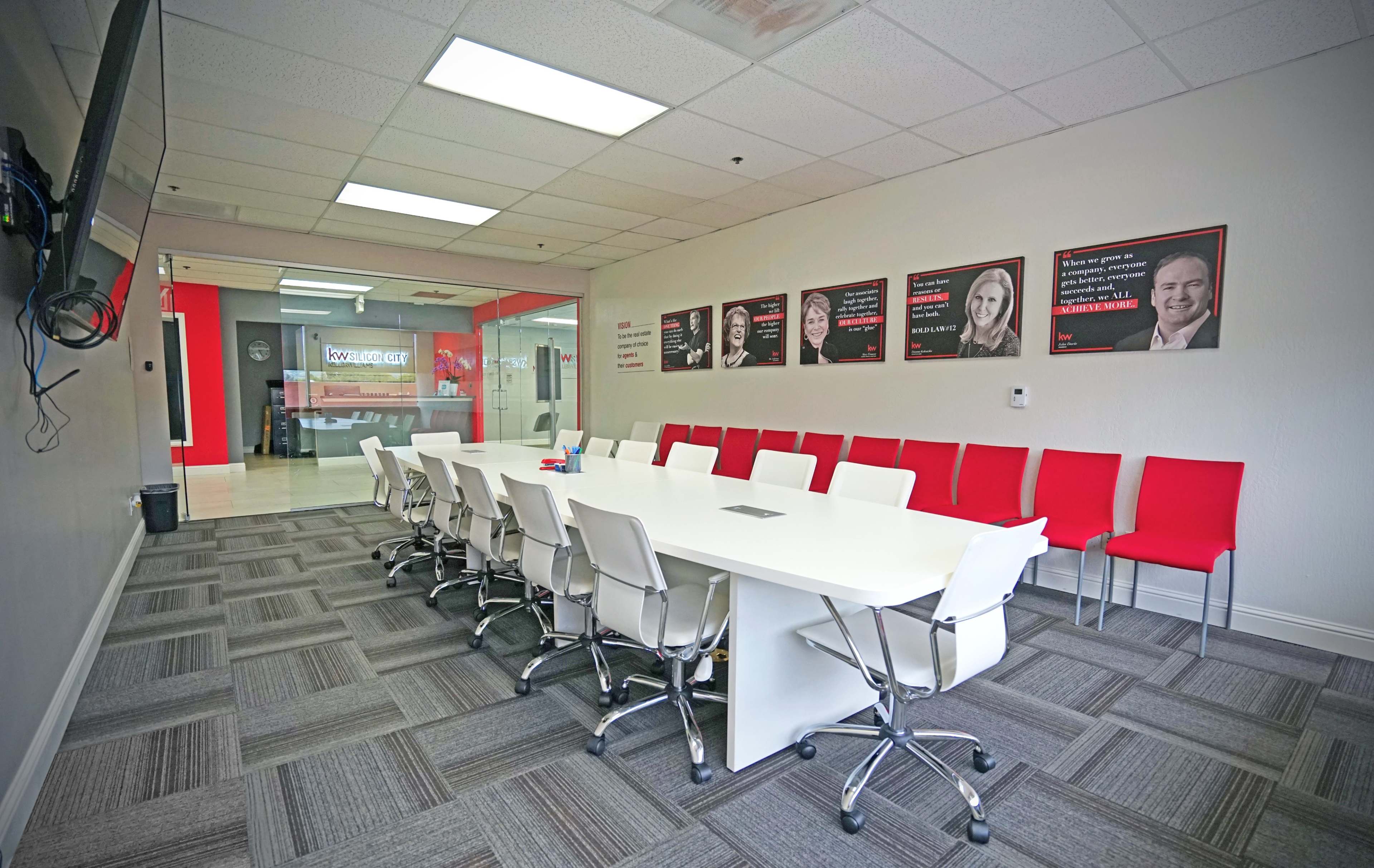 A modern conference room features a long white table surrounded by white chairs and red chairs, with motivational portraits on the walls and a glass partition leading to another workspace.