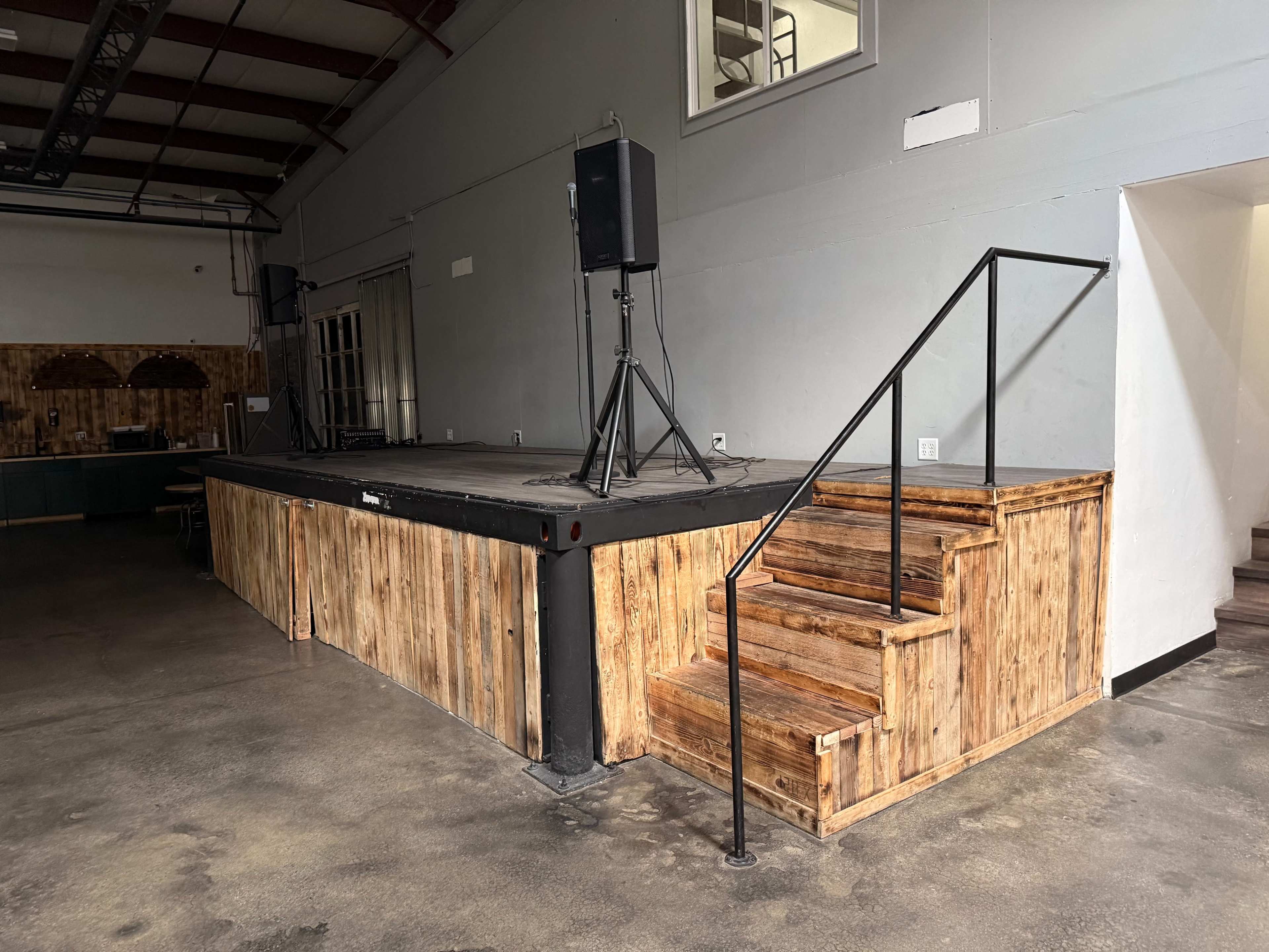 The image shows a wooden stage with a set of stairs leading up to it, surrounded by a plain interior space that includes two speakers on tripods.