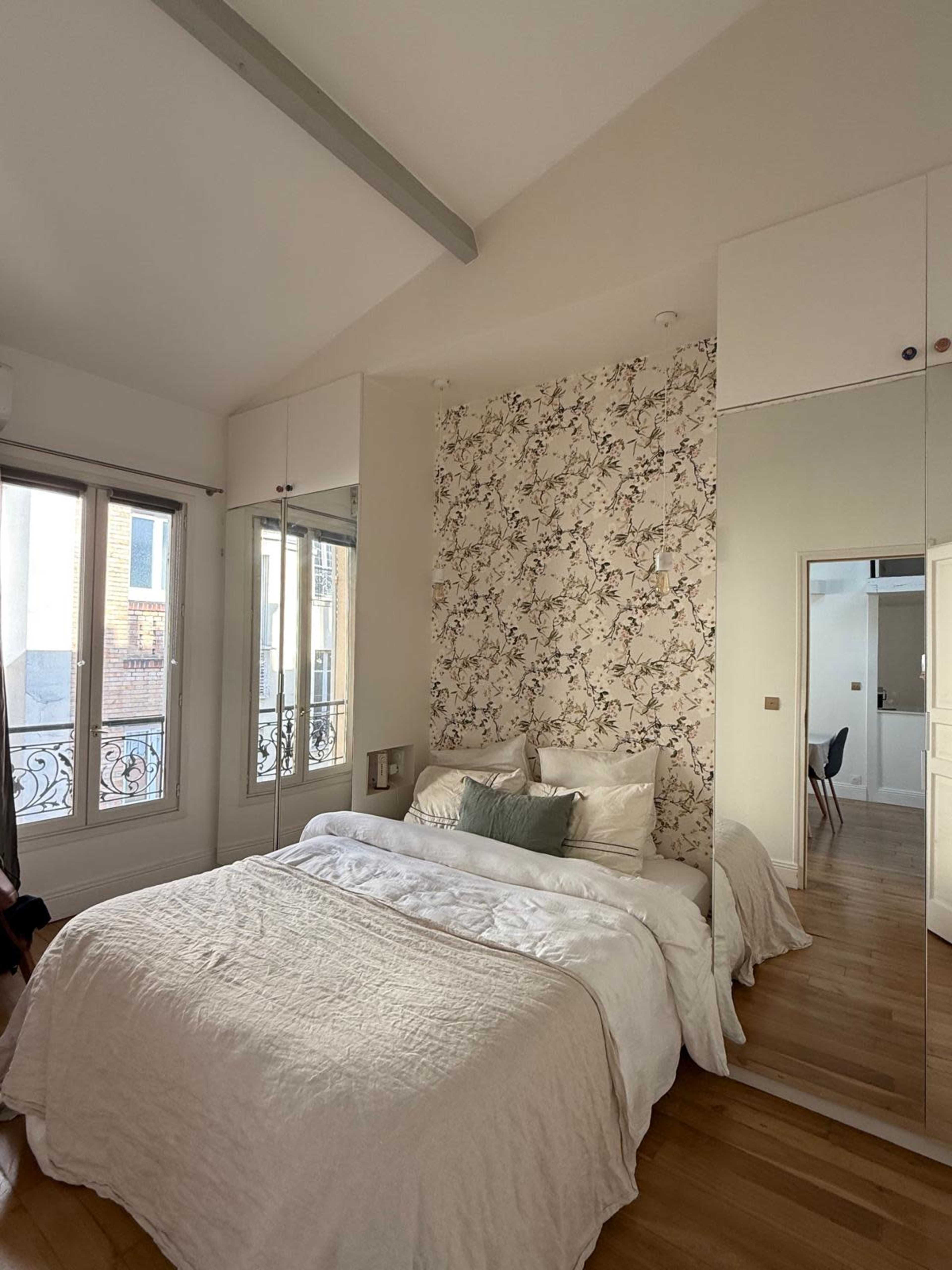 The image shows a bright bedroom featuring a bed with a light-colored duvet, floral wallpaper on one wall, and large mirrors that reflect natural light from the windows.