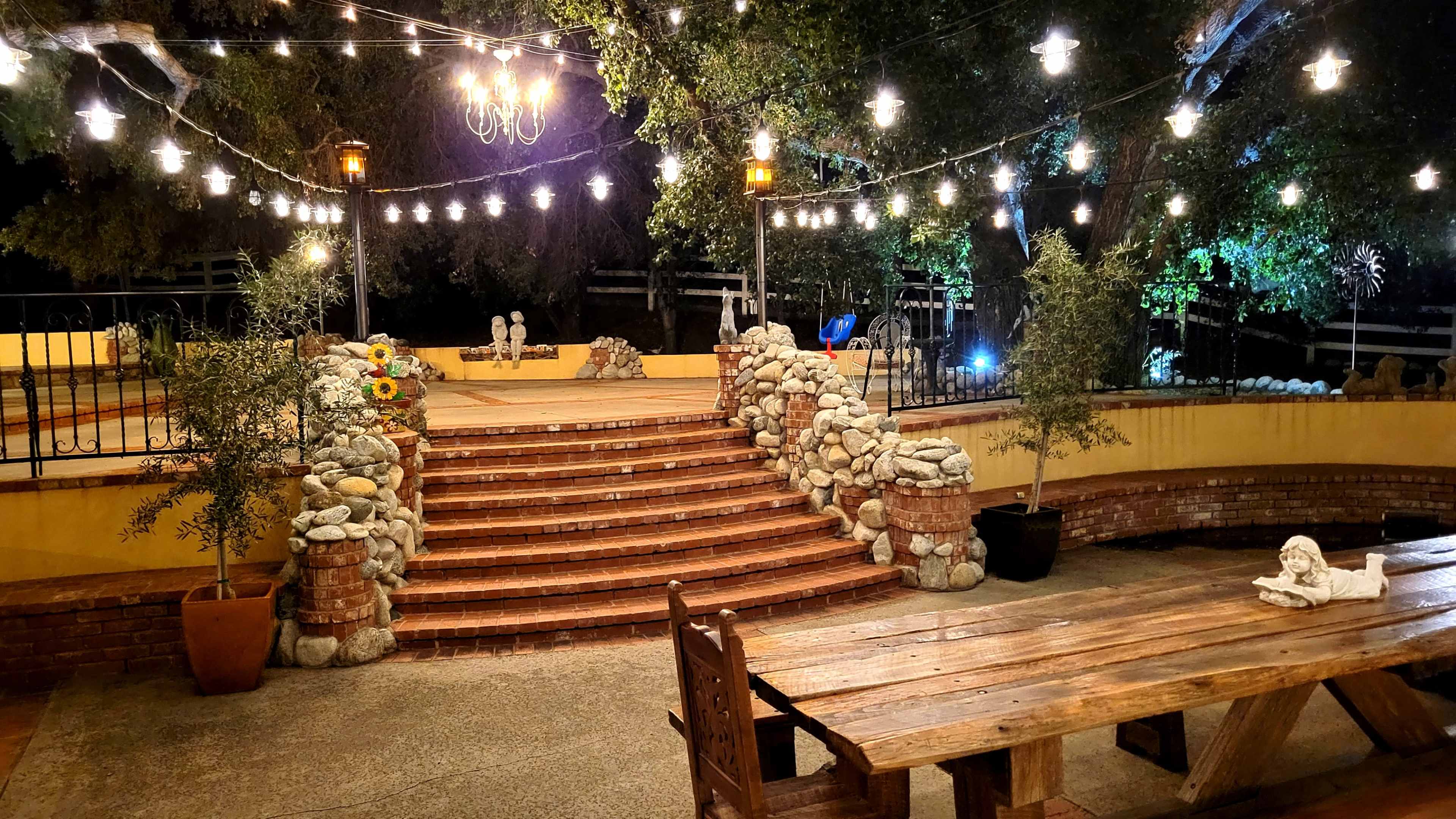 The image depicts an outdoor garden area with stone steps leading to a raised platform, surrounded by string lights and illuminated by lanterns.