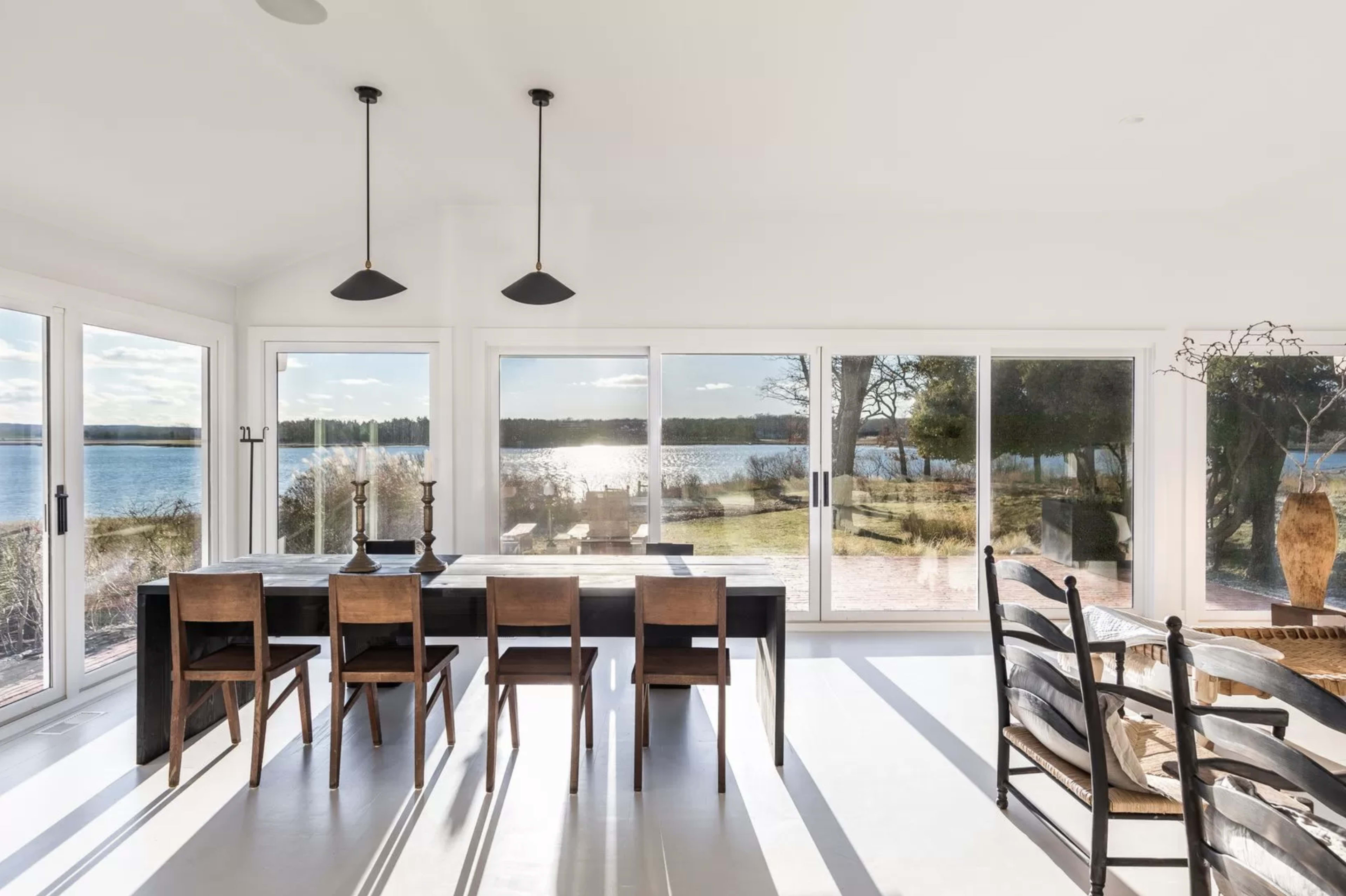 A dining area with a long table and wooden chairs, overlooking a lake through large glass windows.
