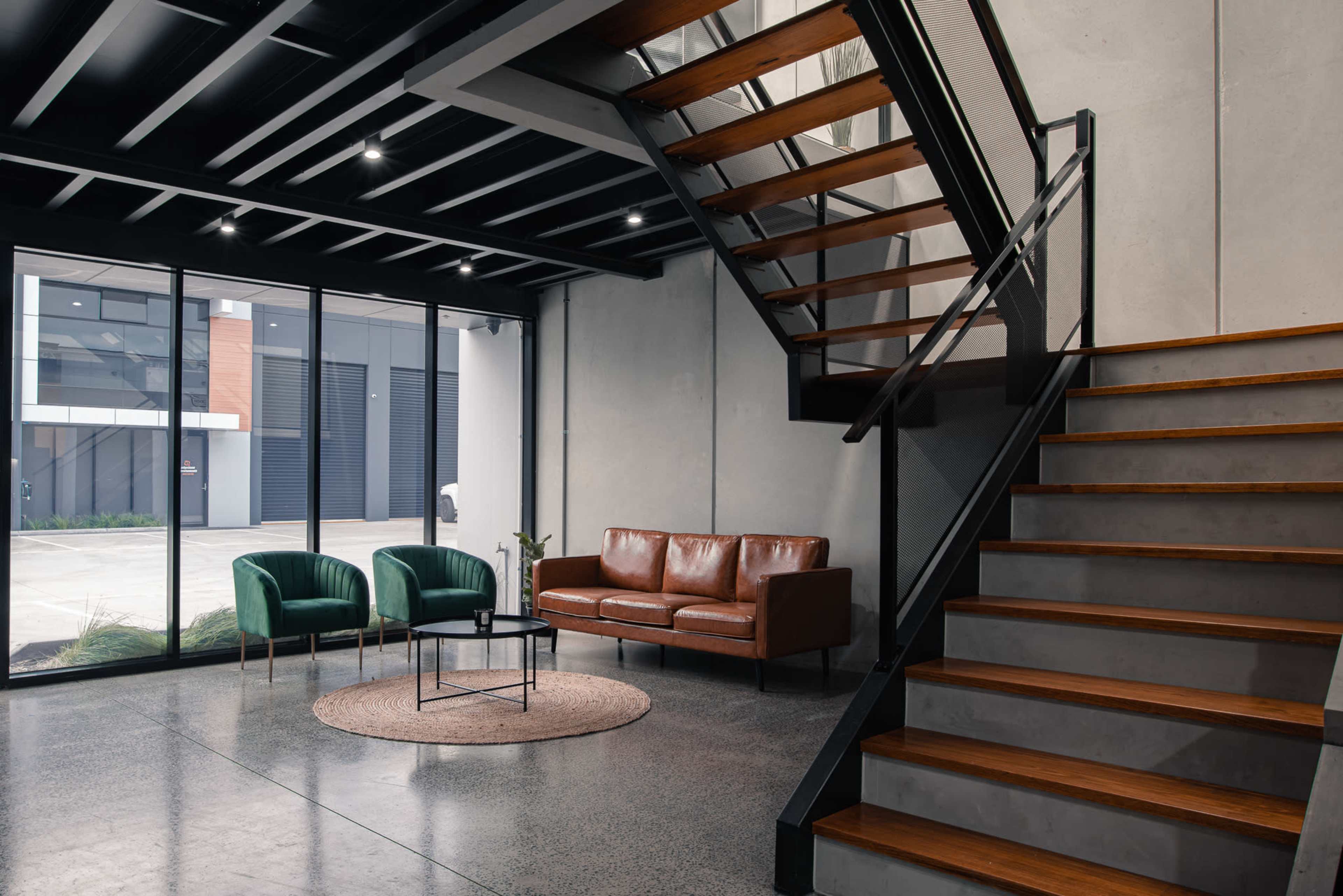 A modern interior with a staircase, a brown leather sofa, two green chairs, and large windows revealing an industrial outdoor view.
