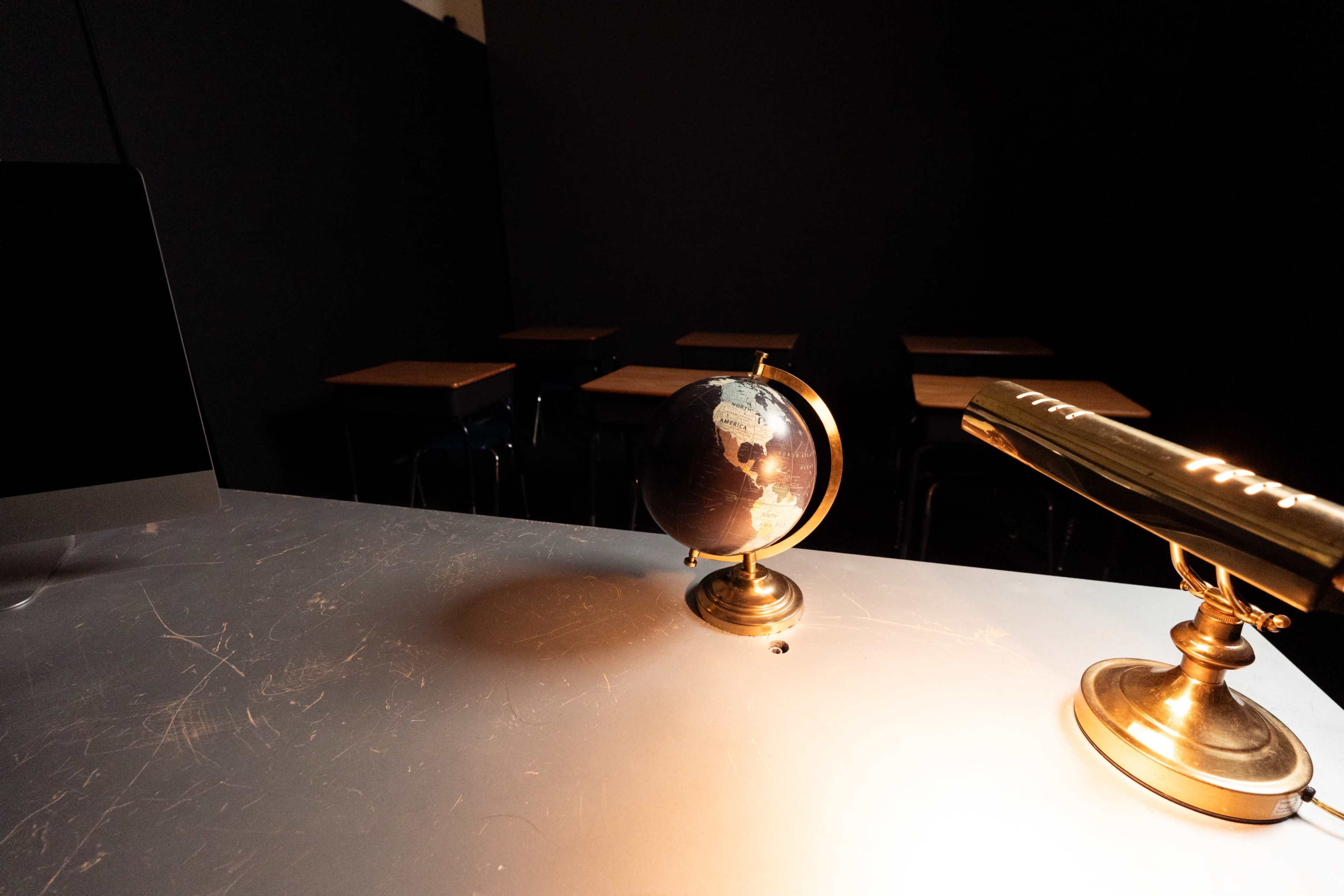 A globe sits on a desk illuminated by a lamp, with empty classroom desks visible in the background.