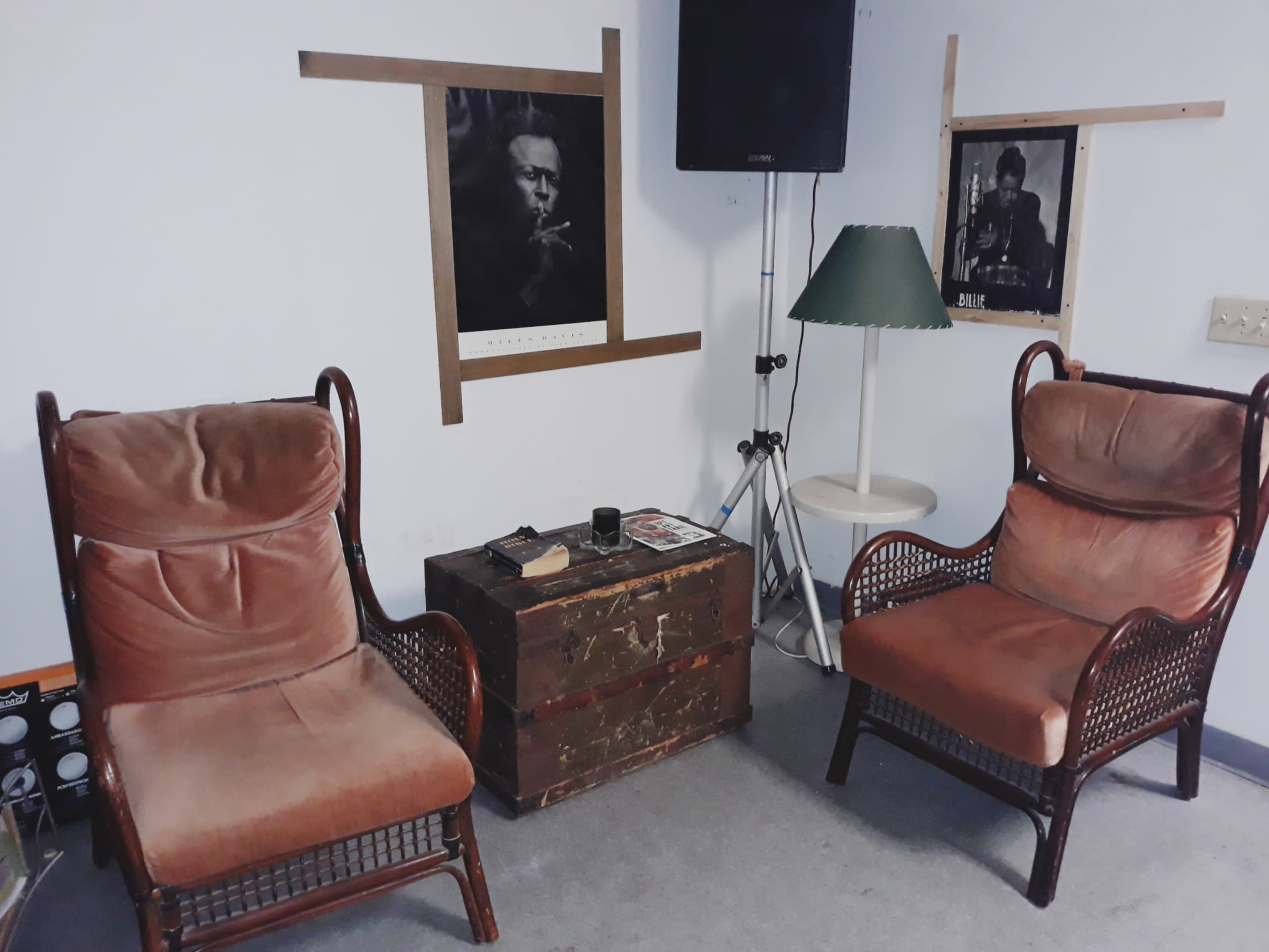Two rattan armchairs facing a wooden chest, with black-and-white posters on the wall and a green lamp nearby.
