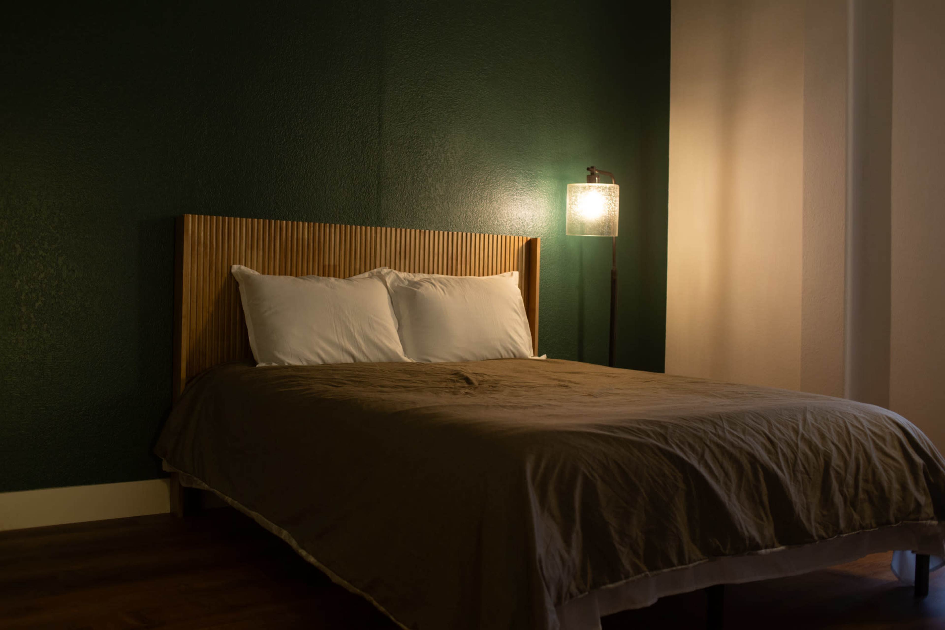 The image shows a simple, modern bedroom with a wooden bed frame, two white pillows, and a lamp beside the bed on a dark green wall.