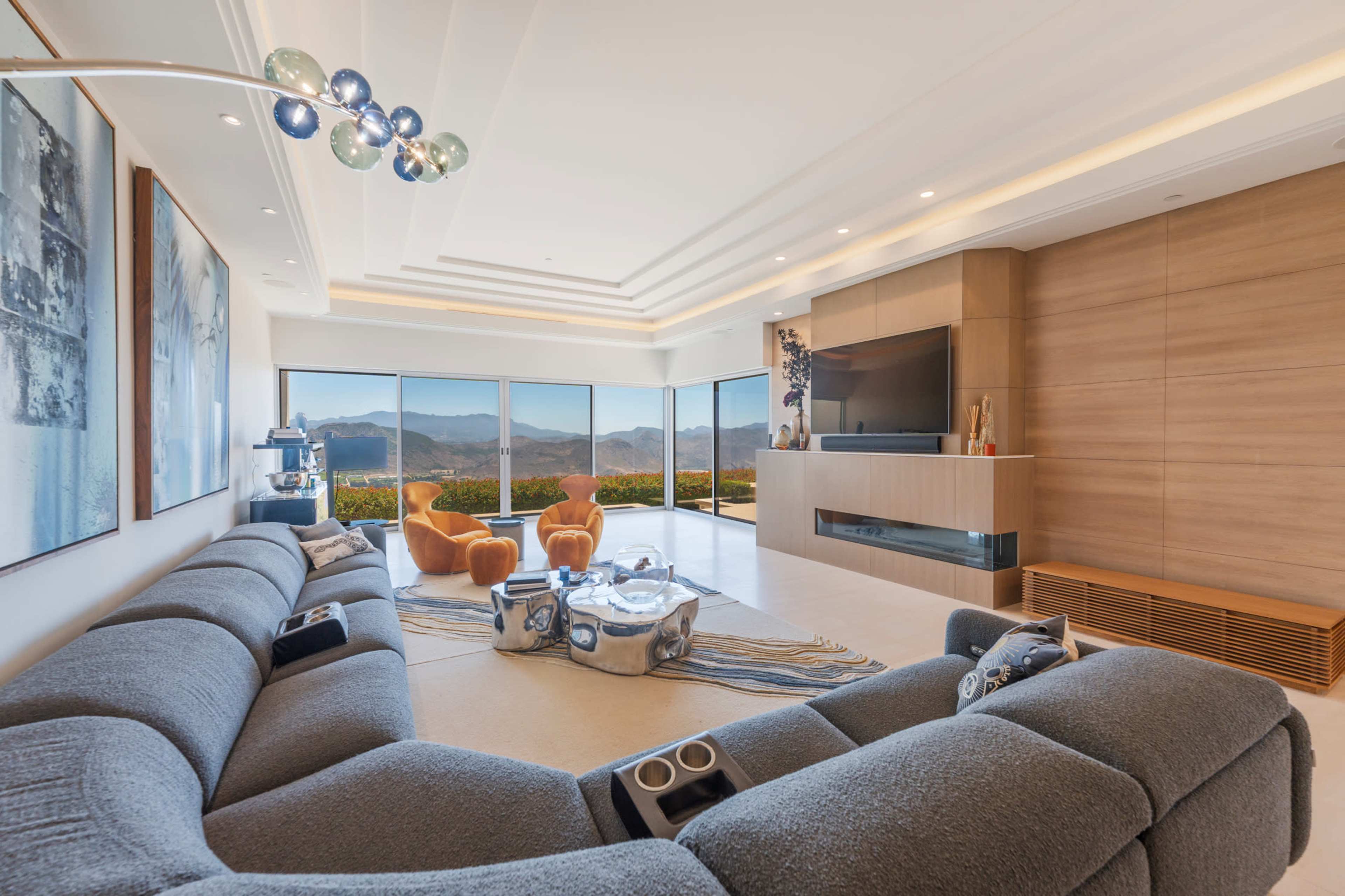 The image shows a modern living room with a large gray sectional sofa, two orange accent chairs, a glass coffee table, and panoramic windows displaying a mountain view.
