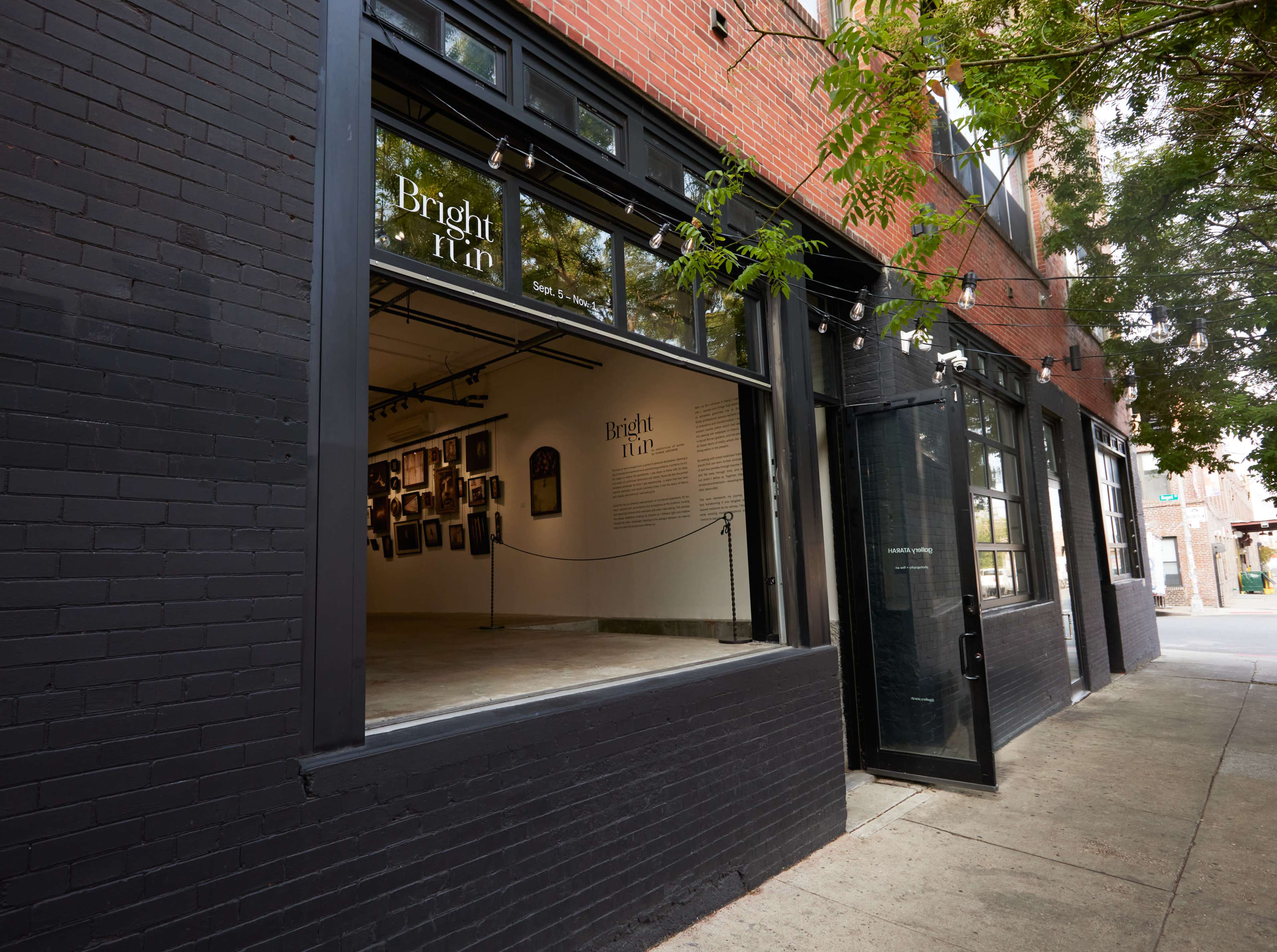 The image shows the exterior of an art gallery with large windows revealing an interior space filled with framed artworks.