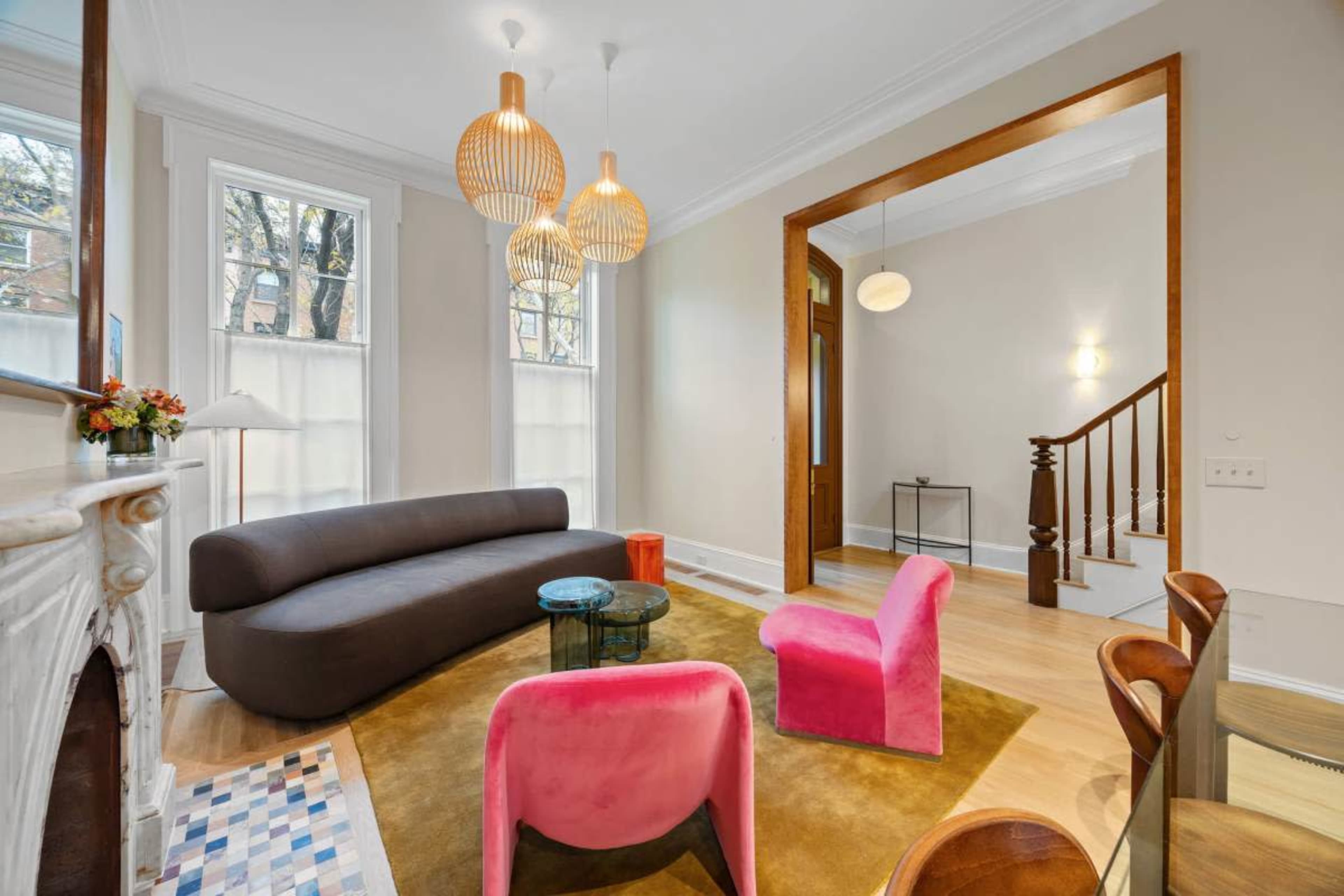The image shows a modern living room featuring a curved gray sofa, two pink chairs, a glass coffee table, and decorative pendant lights, with large windows allowing natural light.