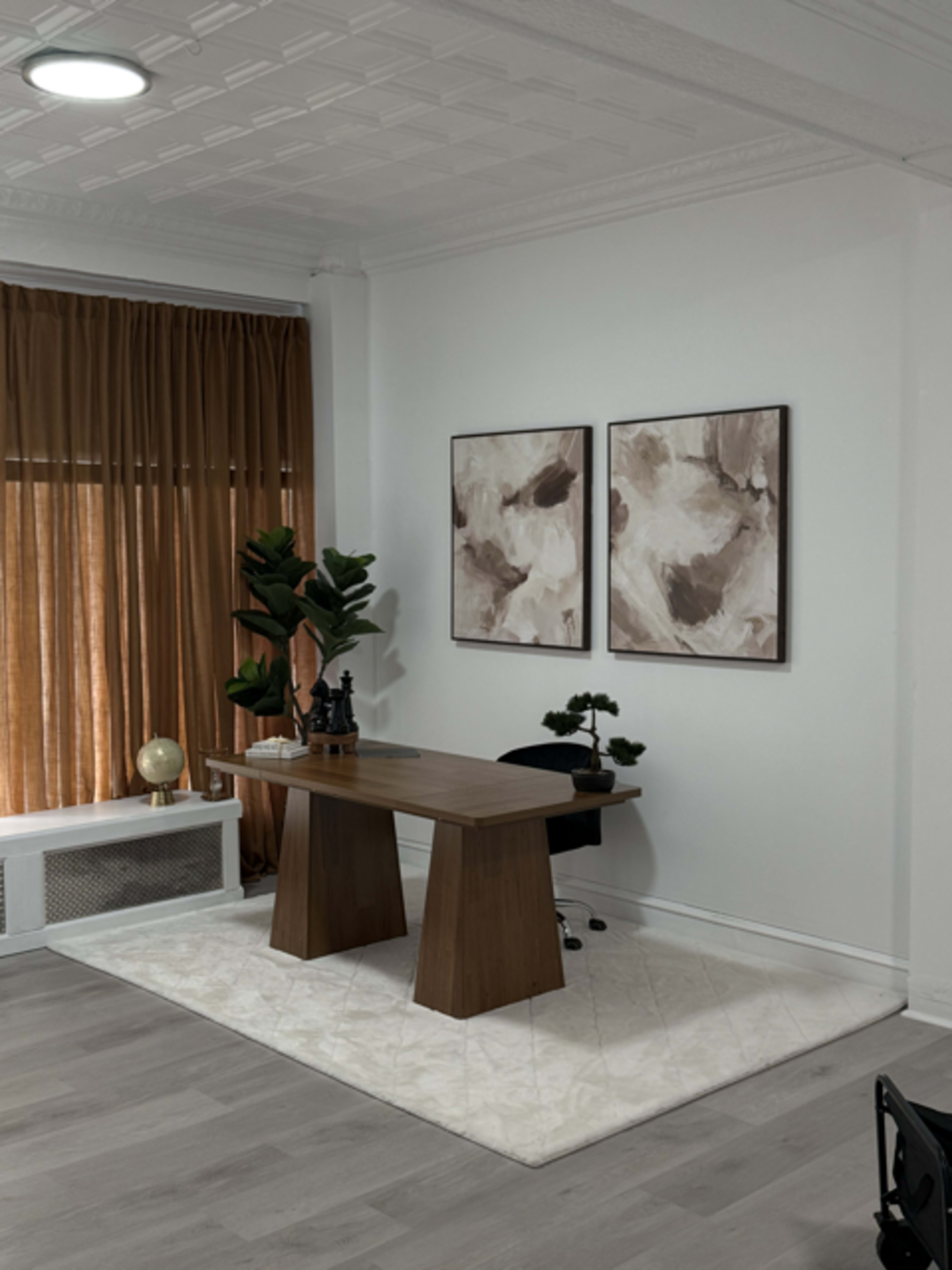 The image shows a modern office space featuring a wooden desk, two abstract art pieces on the wall, a large potted plant, and floor-to-ceiling curtains.