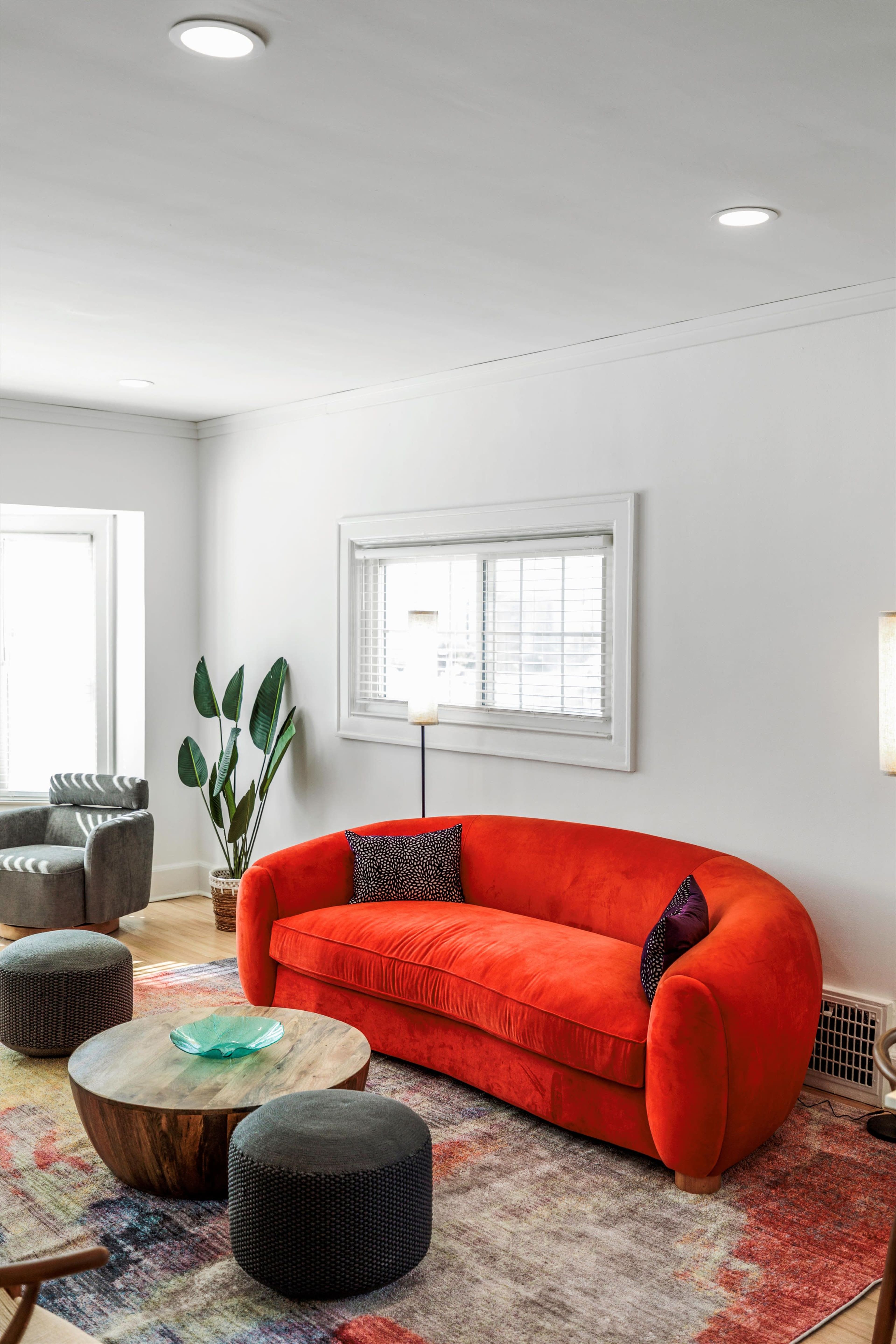 A bright living room features a curved orange sofa, a round wooden coffee table, and patterned ottomans, with natural light streaming through a window.